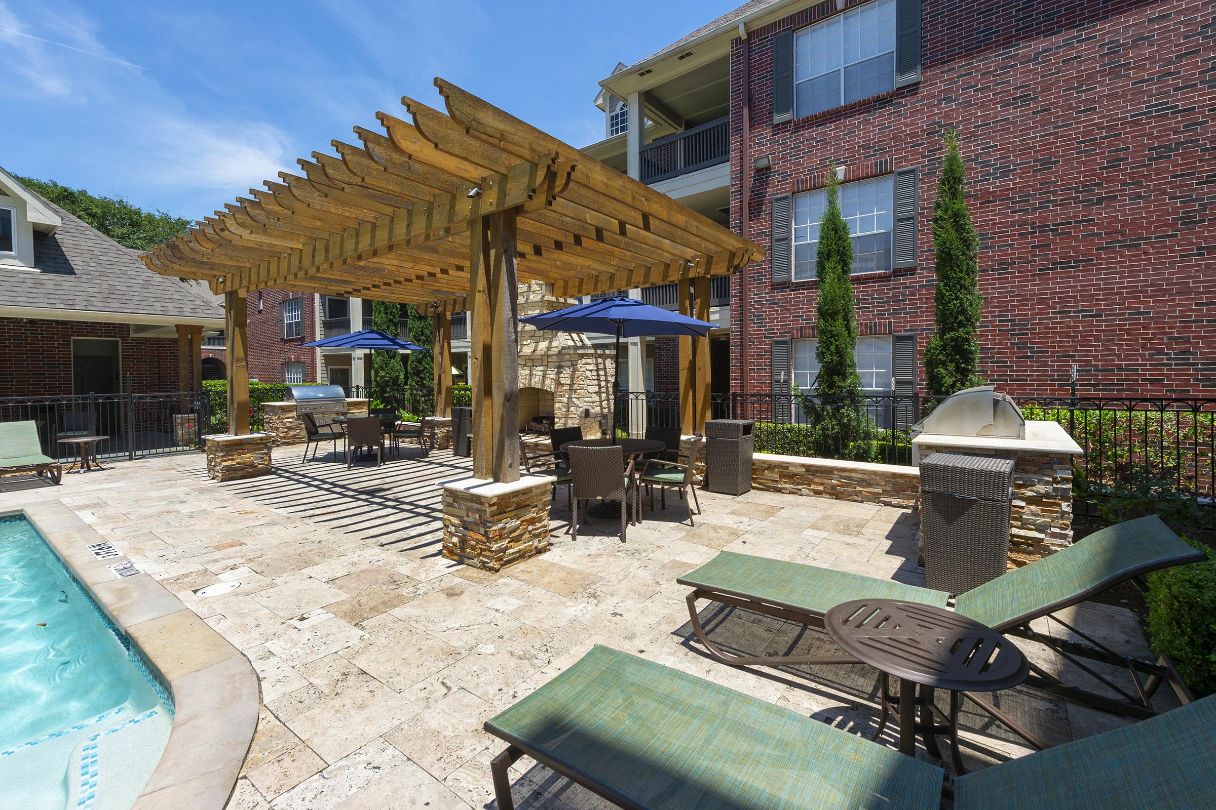 A sunny pool area featuring a stone patio, chaise lounges, and a shaded pergola with seating. There are umbrellas over the tables and lush green landscaping surrounding the space, ideal for relaxation and outdoor gatherings. The backdrop includes a red brick building and clear blue sky.