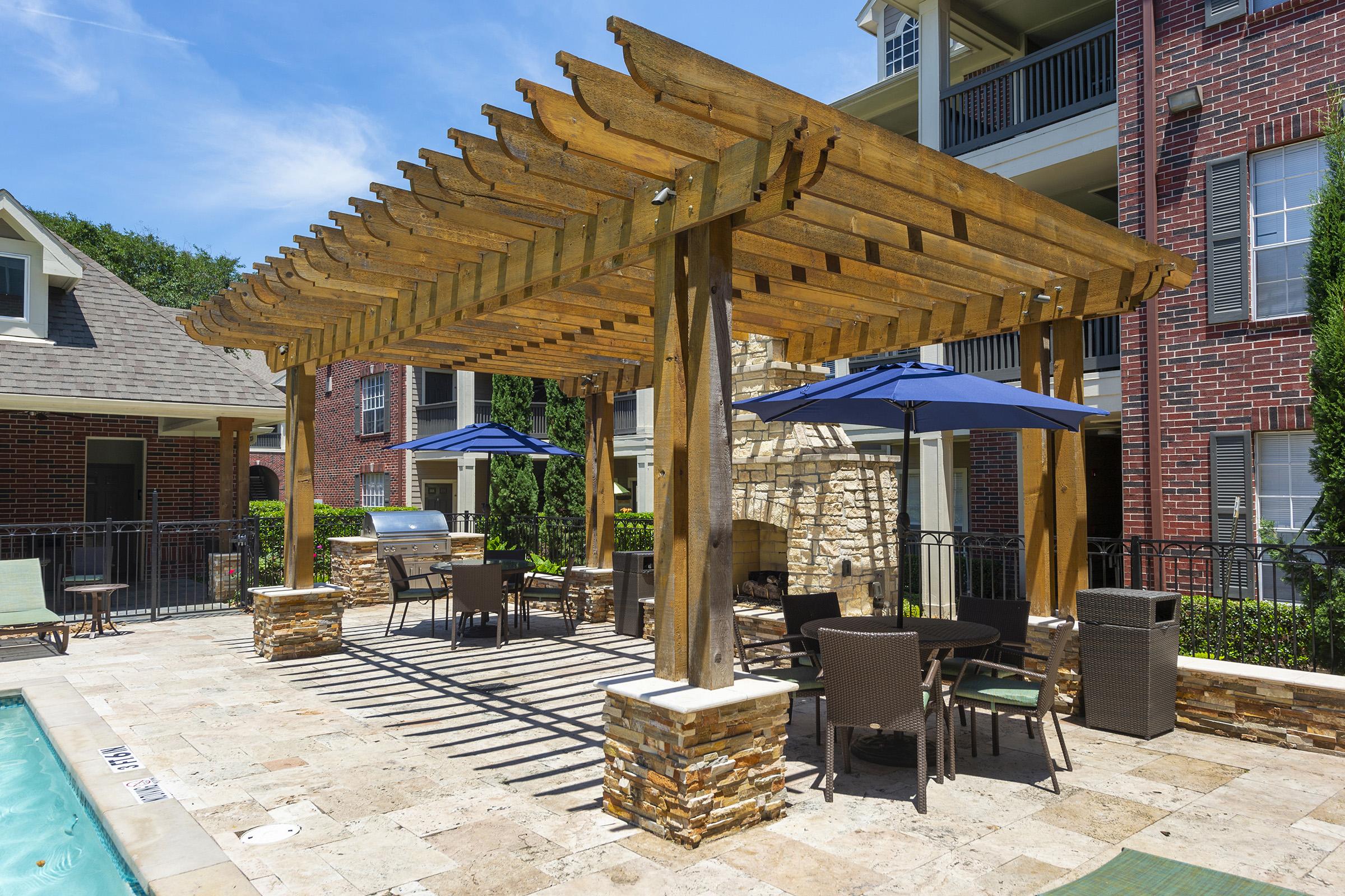 A shaded outdoor seating area with a wooden pergola, featuring patio furniture and umbrellas. Nearby, there's a barbeque grill and a clear pool. The setting includes brick buildings in the background and landscaped greenery, providing a relaxing atmosphere.