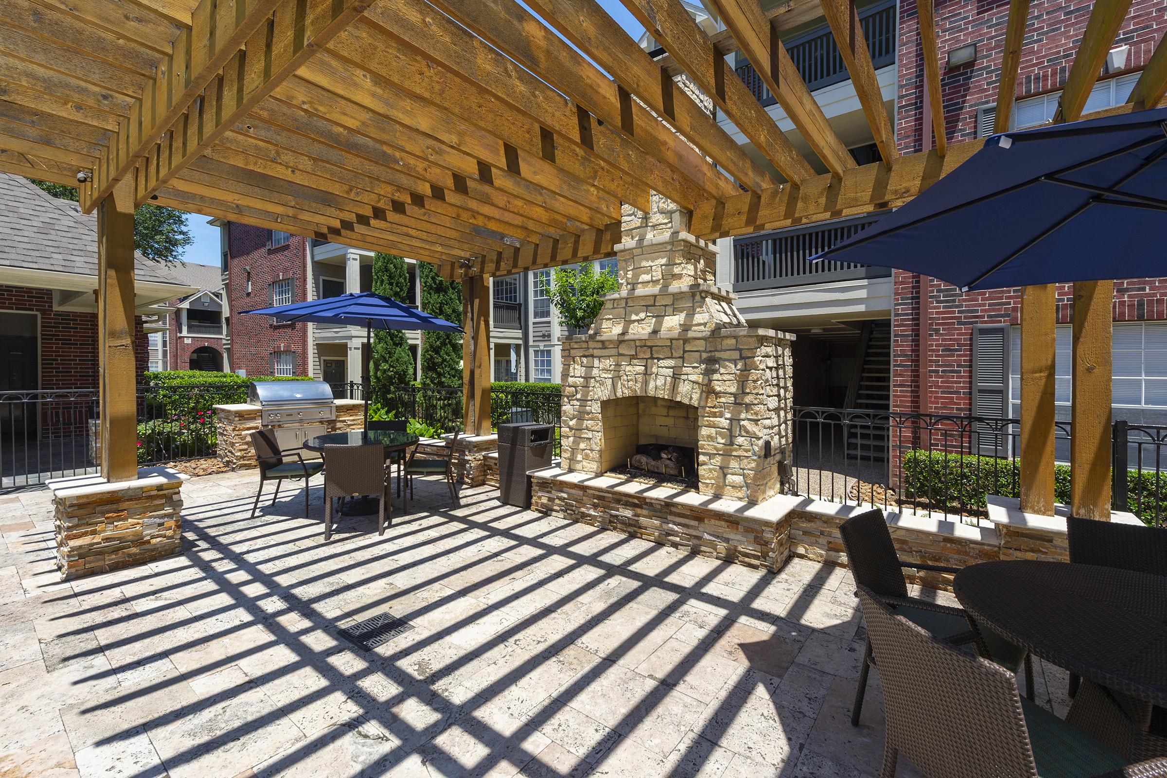 Outdoor seating area with wooden pergola and stone fireplace. Features a grill and shaded tables with umbrellas, surrounded by greenery and brick apartment buildings. Bright sunlight creates striped shadows on the paved floor.