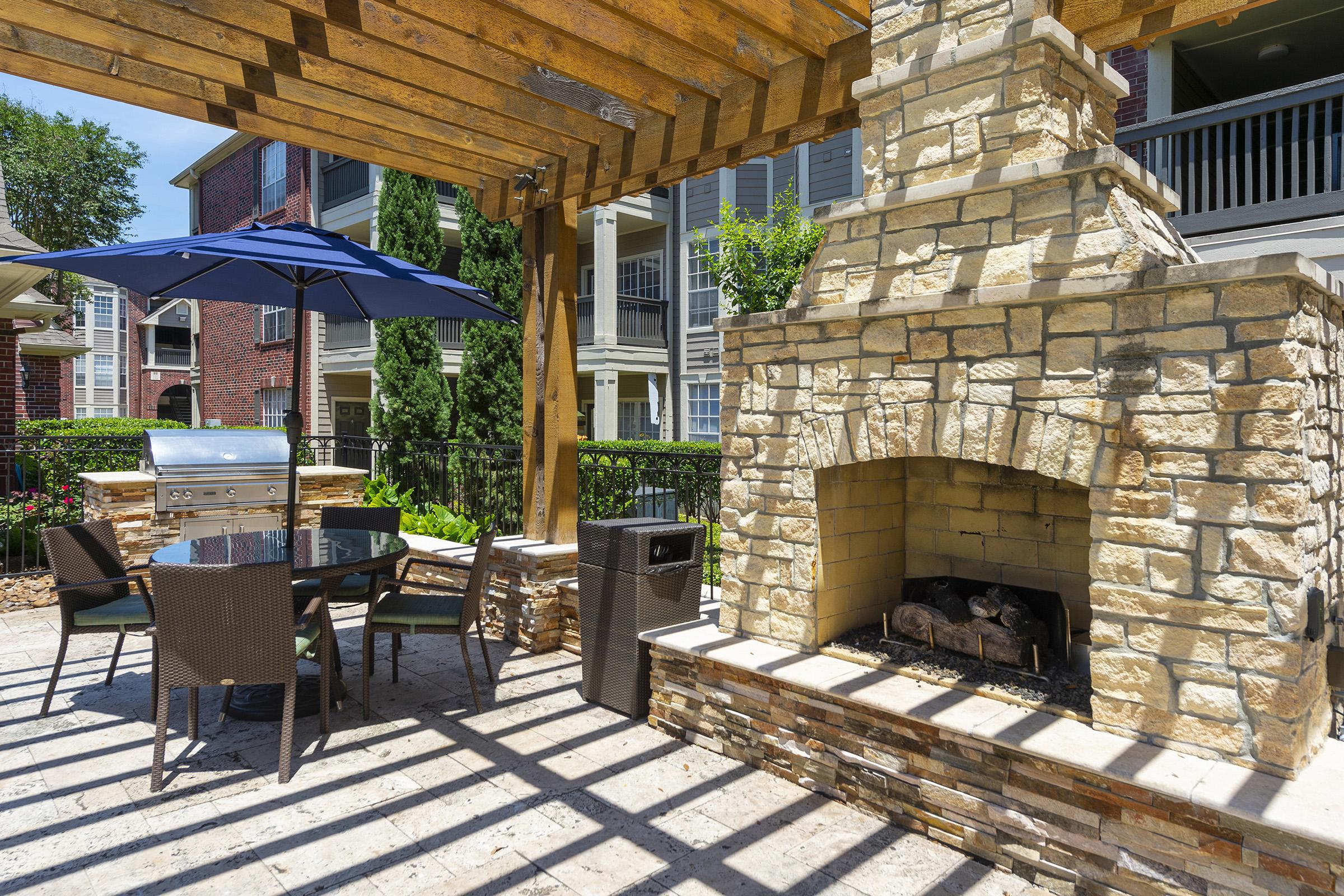 Outdoor patio area with a stone fireplace, grill, and a table with chairs under a wooden pergola. An umbrella provides shade, and lush greenery surrounds the space, enhancing the inviting atmosphere.