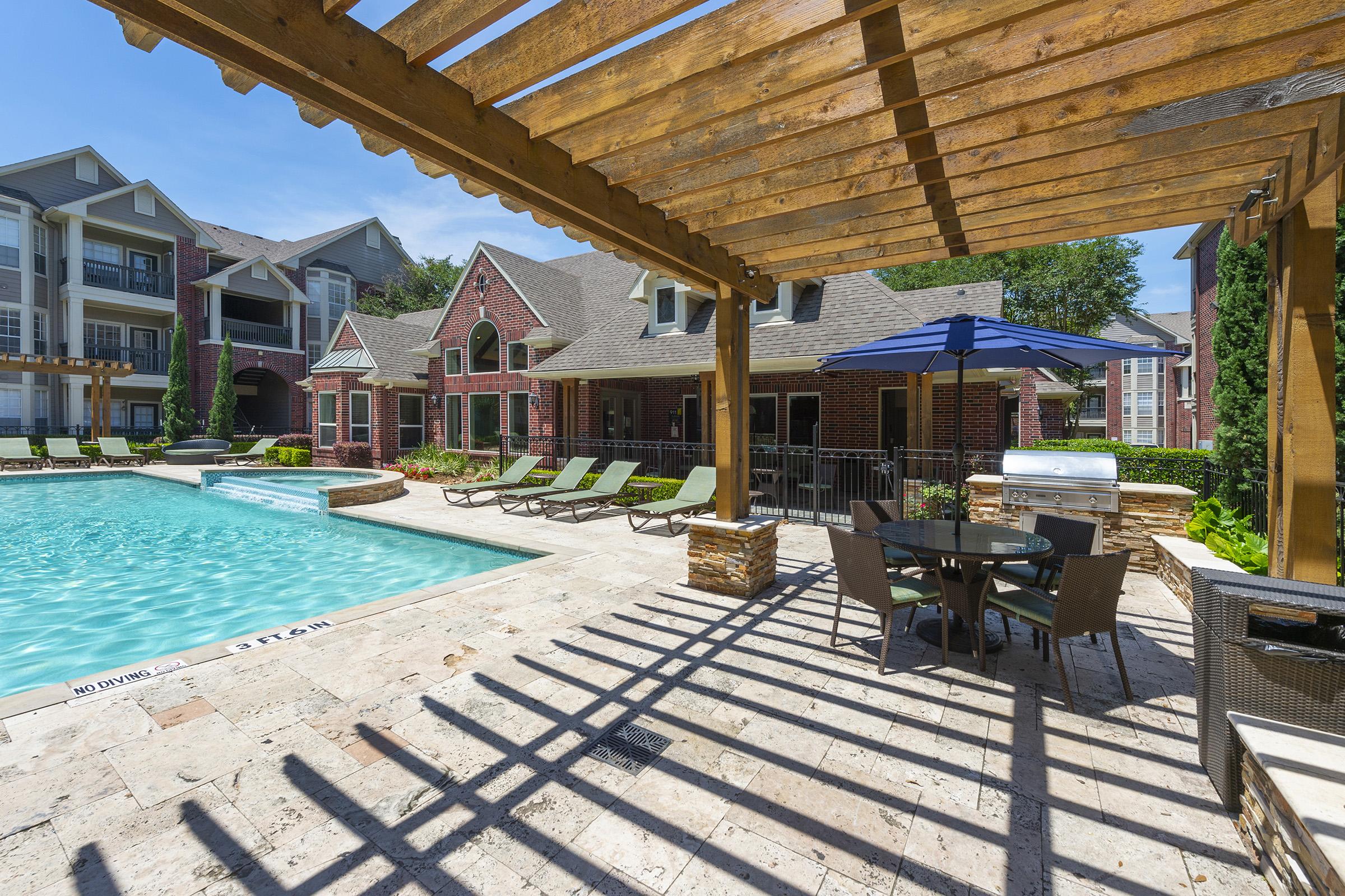 A bright outdoor swimming pool surrounded by a stone patio, sun loungers, and a shaded dining area under a wooden pergola. In the background, stylish apartment buildings can be seen, with a clear blue sky overhead.