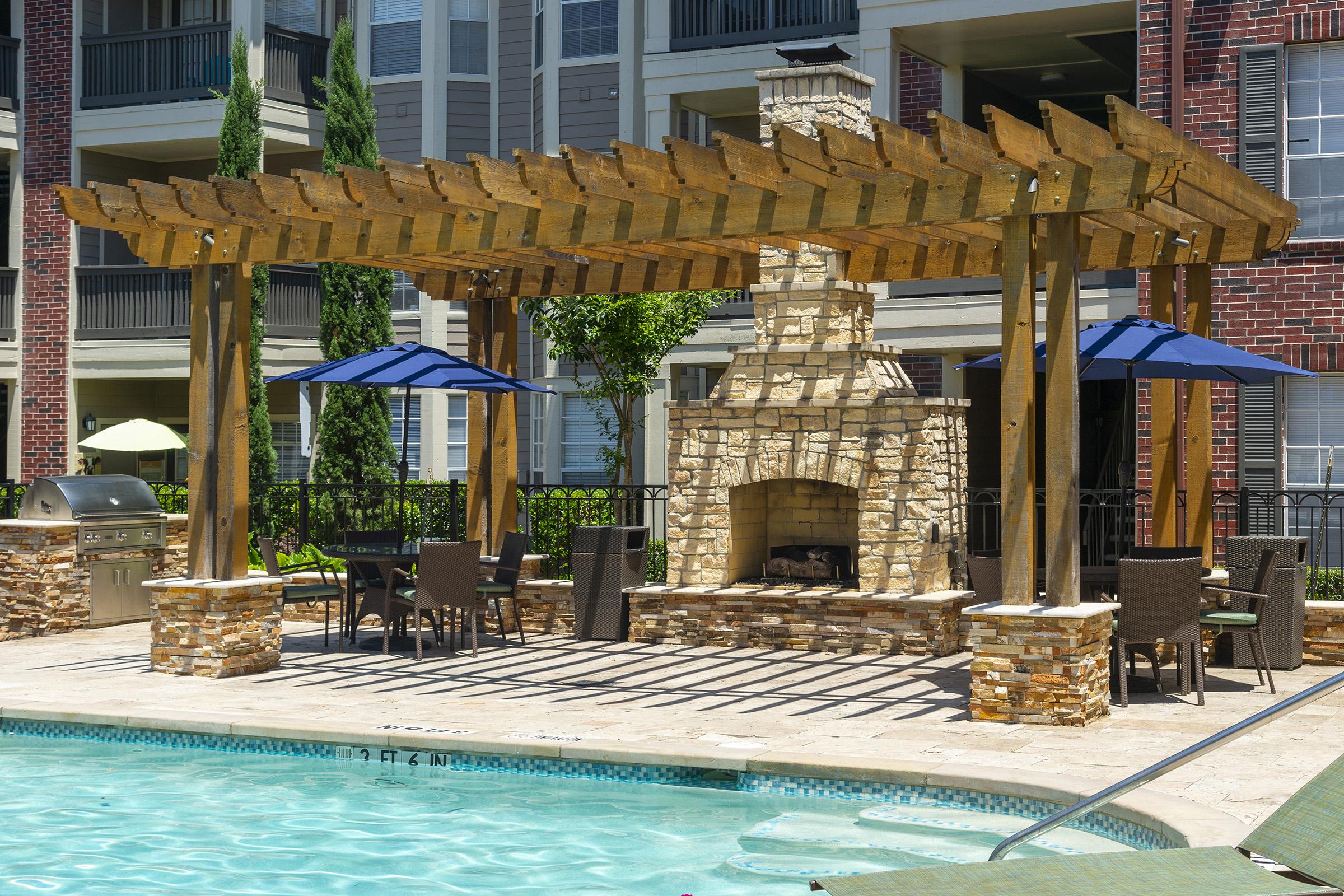 A poolside area featuring a wooden pergola with a stone fireplace underneath. The space includes a set of outdoor tables and chairs, two blue umbrellas for shade, a gas grill, and a clear swimming pool surrounded by landscaped greenery and apartment buildings in the background.