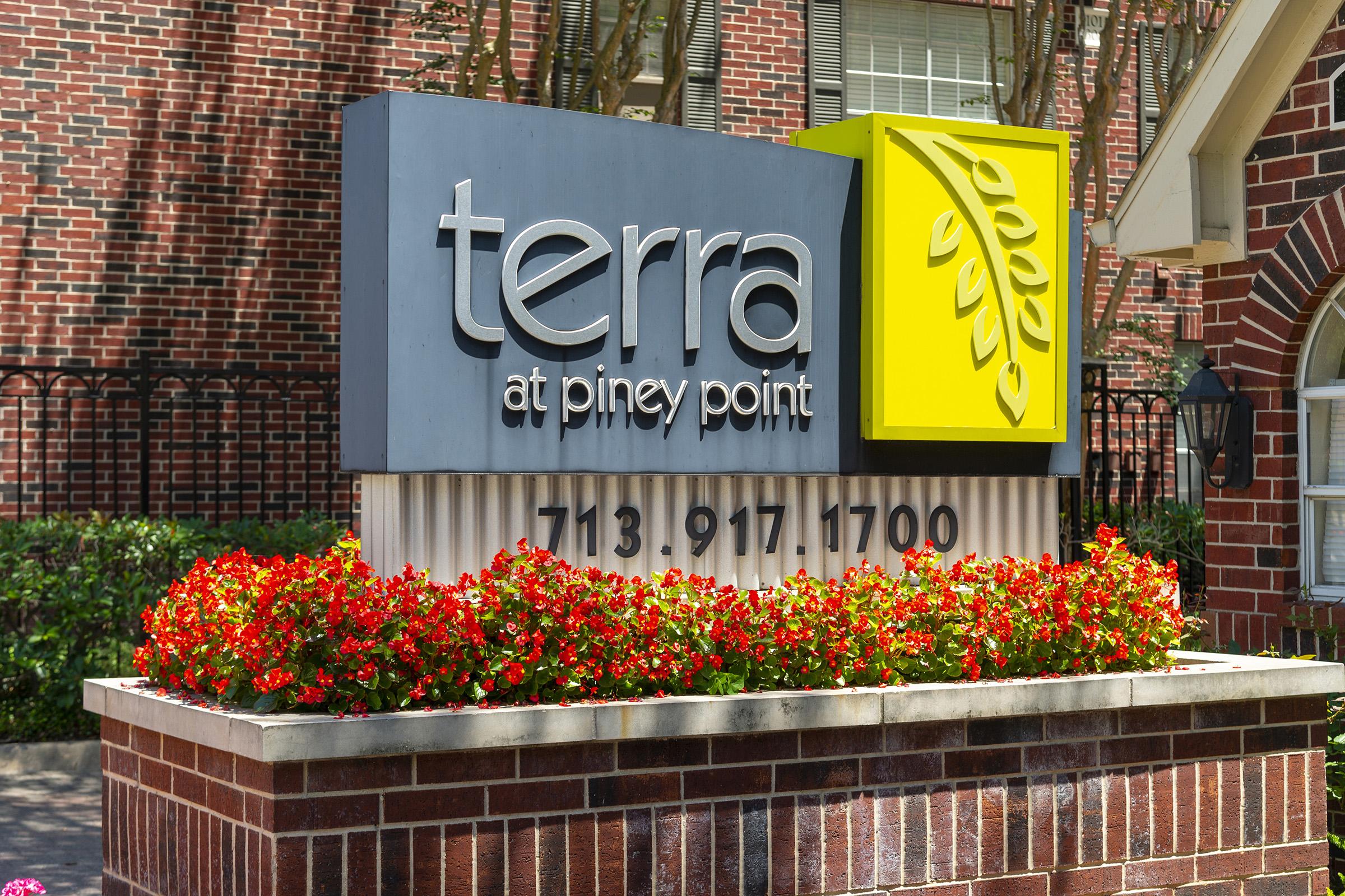 Sign for "terra at piney point" featuring a modern design with a yellow leaf graphic, surrounded by vibrant red flowers in a planter. Contact number displayed: 713-917-1700. The background shows a brick building and greenery.
