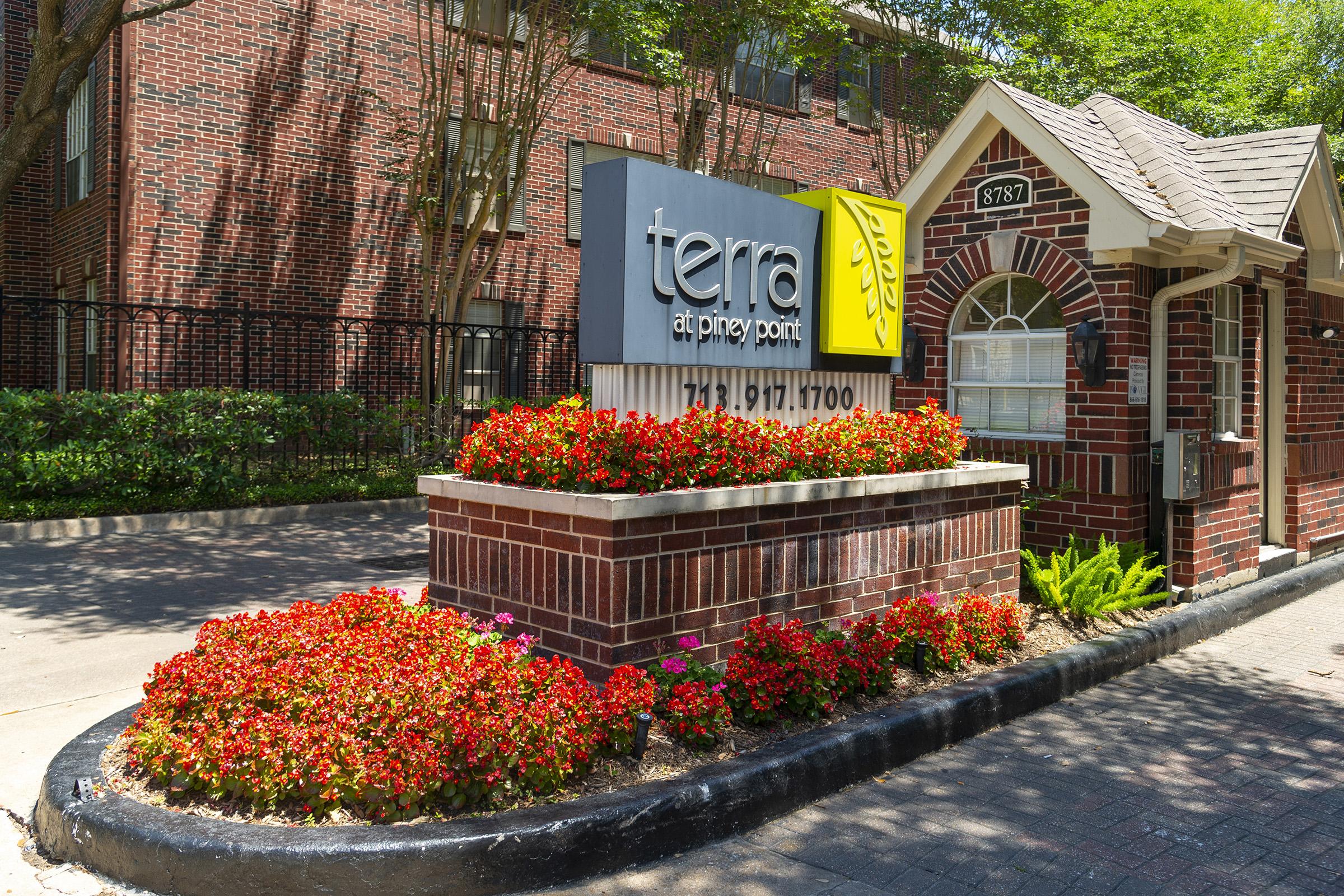 Sign for "terra at piney point" featuring a large logo and contact number, surrounded by vibrant red and orange flowers. The entrance is framed by brick architecture, with green foliage in the background, showcasing a well-maintained landscape in a sunny outdoor setting.
