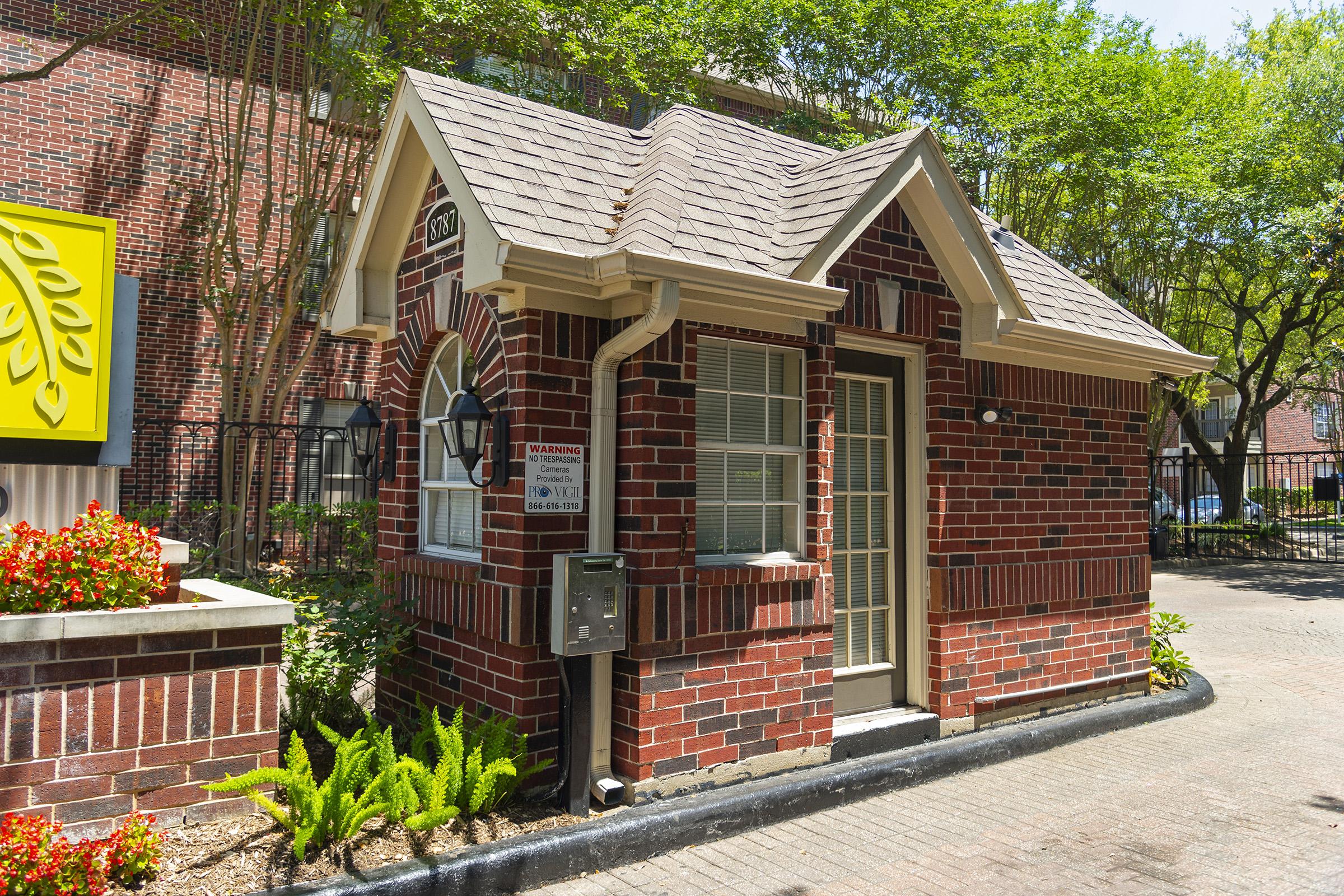A small brick building with a pitched roof, featuring large windows and a door. The structure has decorative brickwork and is surrounded by greenery and flowers. It appears to be a guardhouse or information kiosk, located near a paved walkway. Bright sunlight enhances the colors of the bricks and plants.
