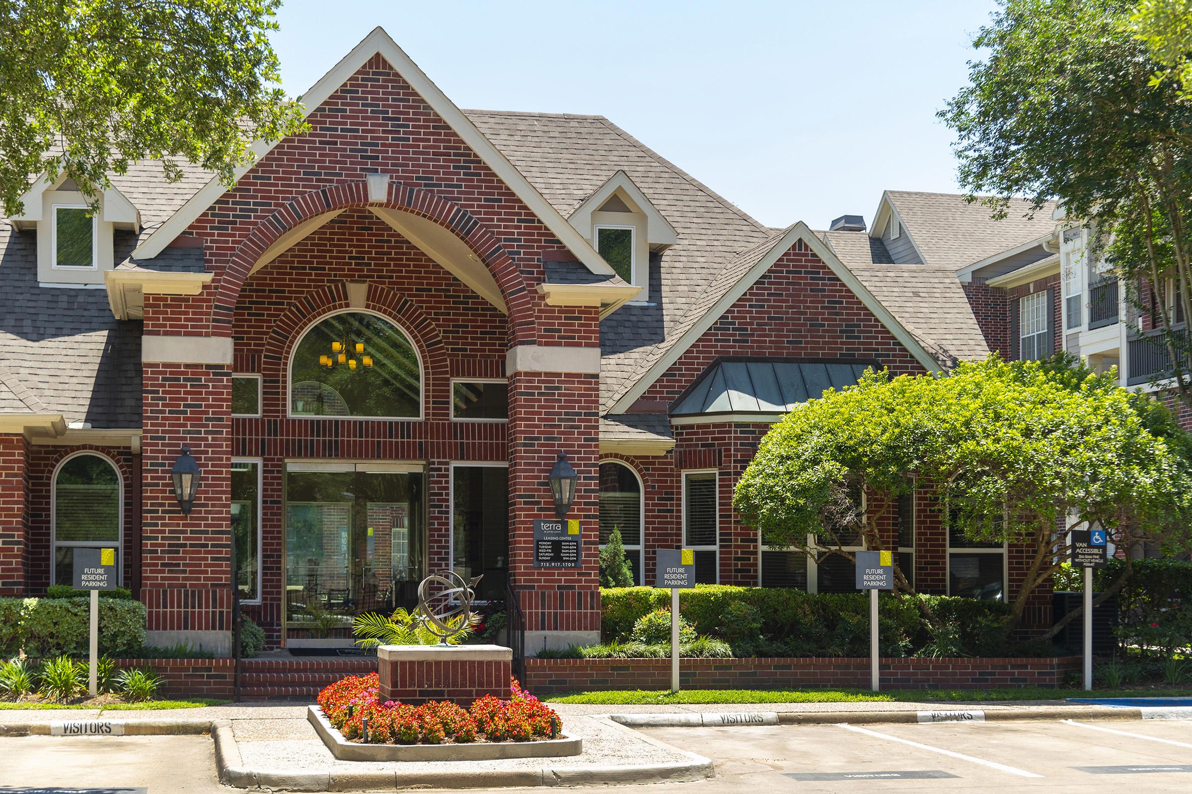 A well-maintained brick building with a large archway entrance, surrounded by vibrant green trees and colorful flower beds. The exterior features multiple gables and large windows, creating an inviting atmosphere in a sunny environment. Signs are visible near the entrance.