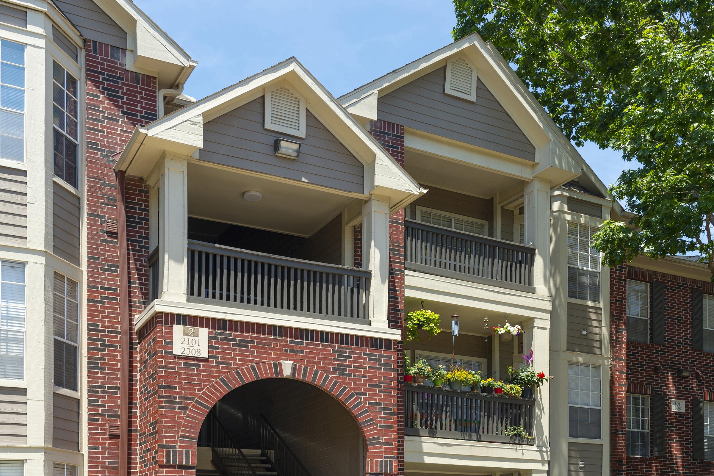 A close-up view of a residential apartment building showcasing a brick facade and wooden balconies. The upper unit features potted flowers on the balcony, while the lower level has a staircase leading to the entrance. Trees provide shade in the foreground under a clear blue sky.