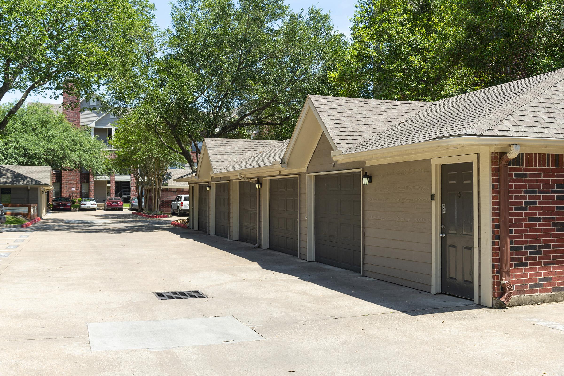 A row of garages with beige doors and a brick wall, situated in a well-maintained residential area. The scene features green trees providing shade, with parked cars visible in the background. Bright sunlight casts shadows on the paved driveway, creating a welcoming atmosphere.