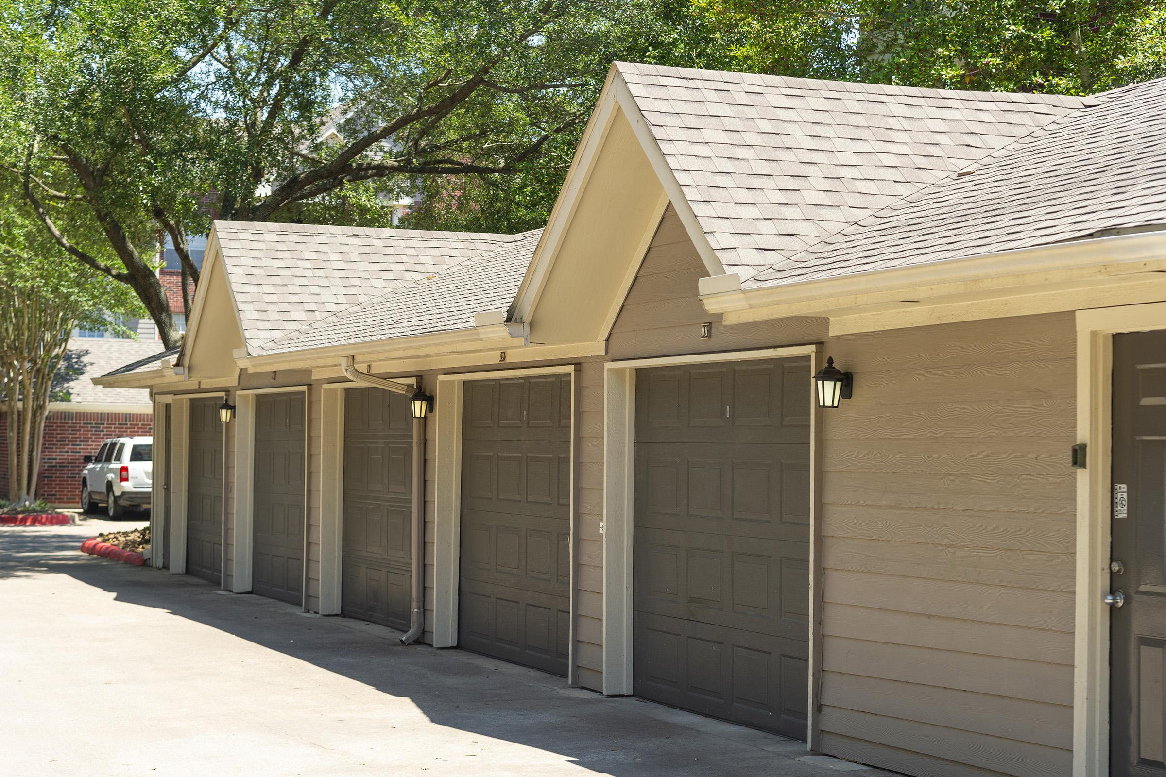 Row of garages with light brown siding and grey doors, set in a paved area surrounded by greenery. Trees provide shade, creating a pleasant, serene environment. The garages have uniform roofs with visible shingles and outdoor lighting fixtures on the sides.