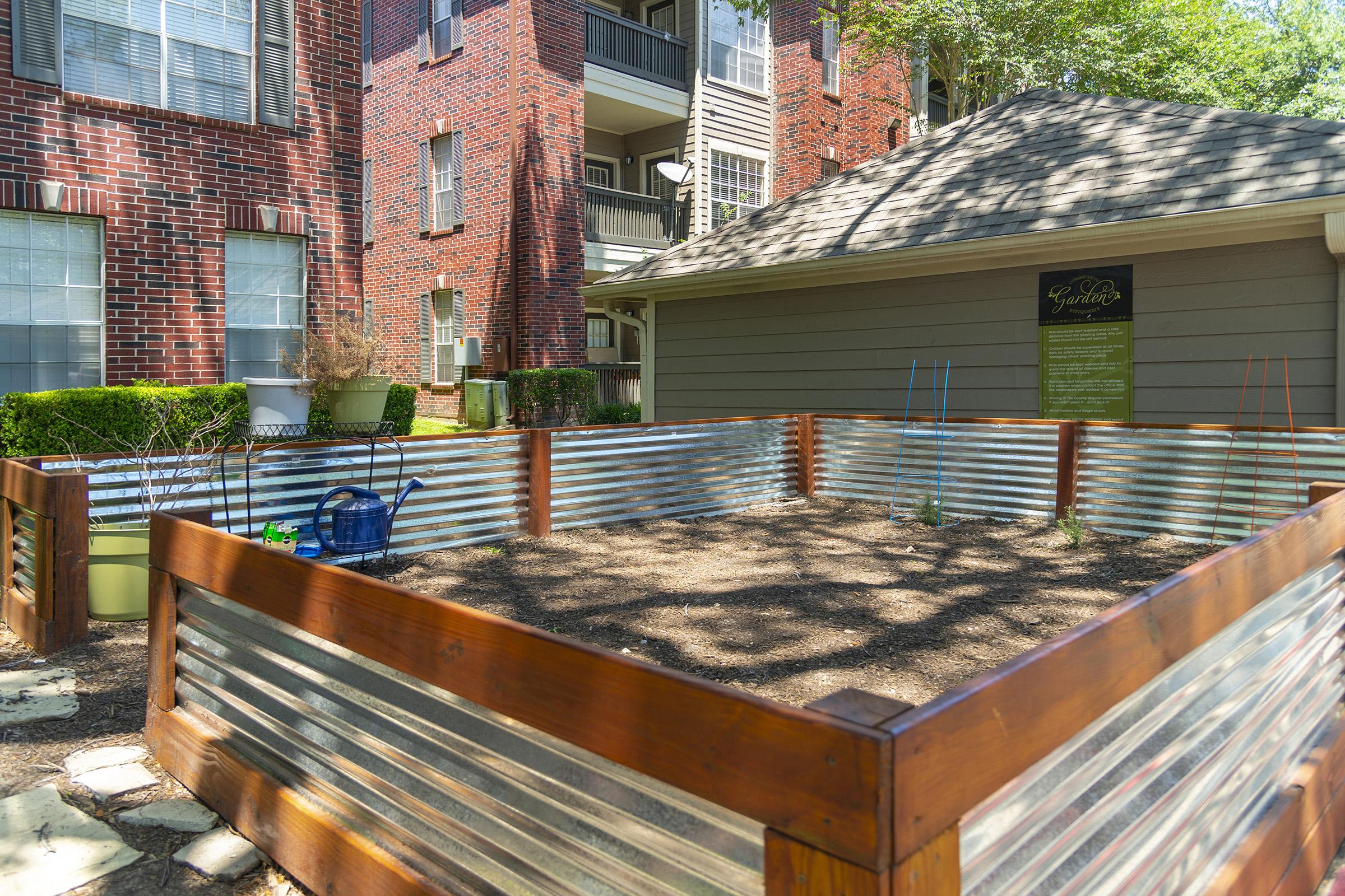 A community garden space with a wooden and metal fence, located next to an apartment building. The area is empty with soil prepared for planting, surrounded by greenery. A nearby sign provides information about the garden. Large windows of the building are visible in the background.