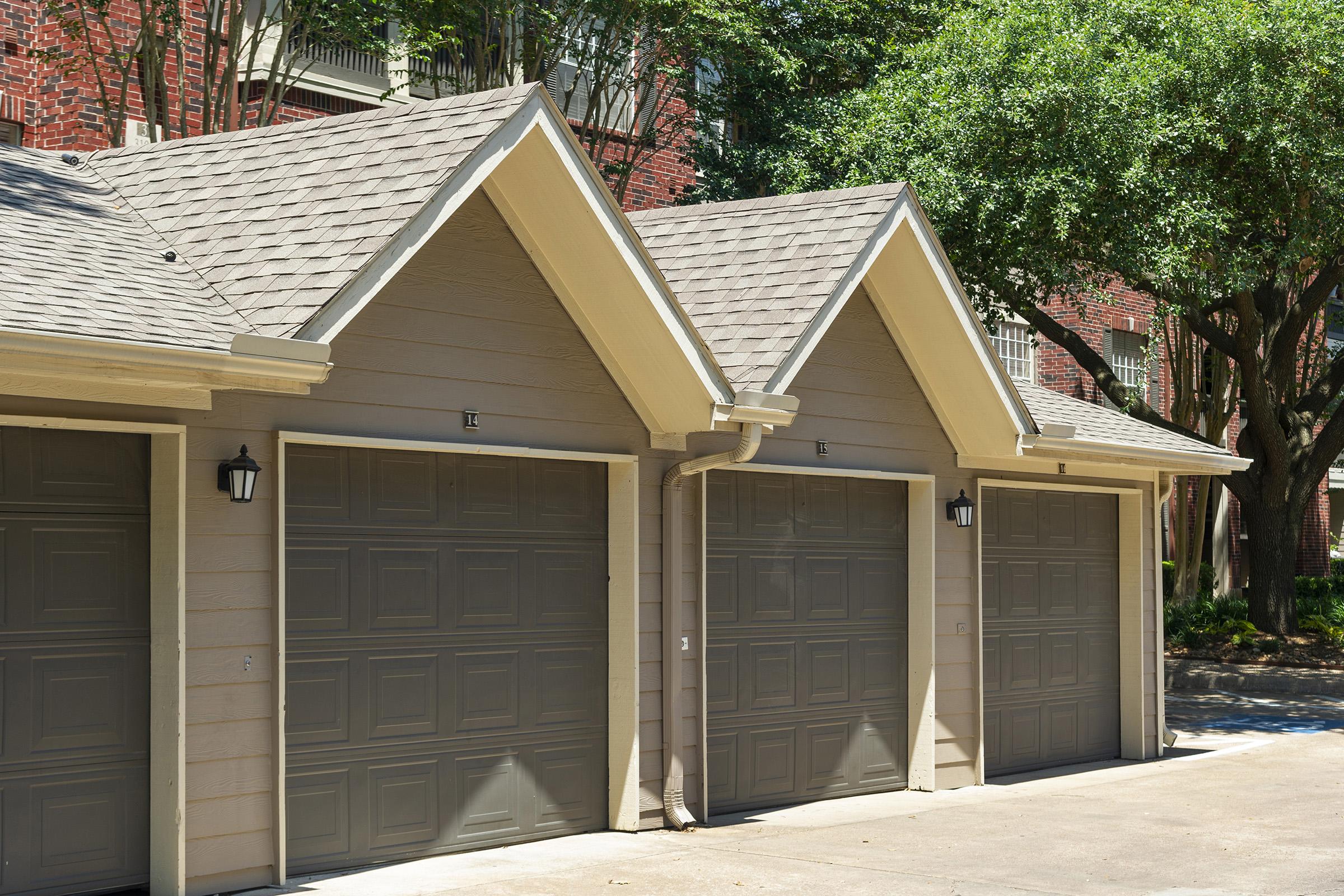 A row of three residential garage doors with light gray panels, featuring peaked roofs and outdoor light fixtures. The garages are situated in a shaded area surrounded by green trees and brick buildings in the background. The driveway is clear and landscaped.