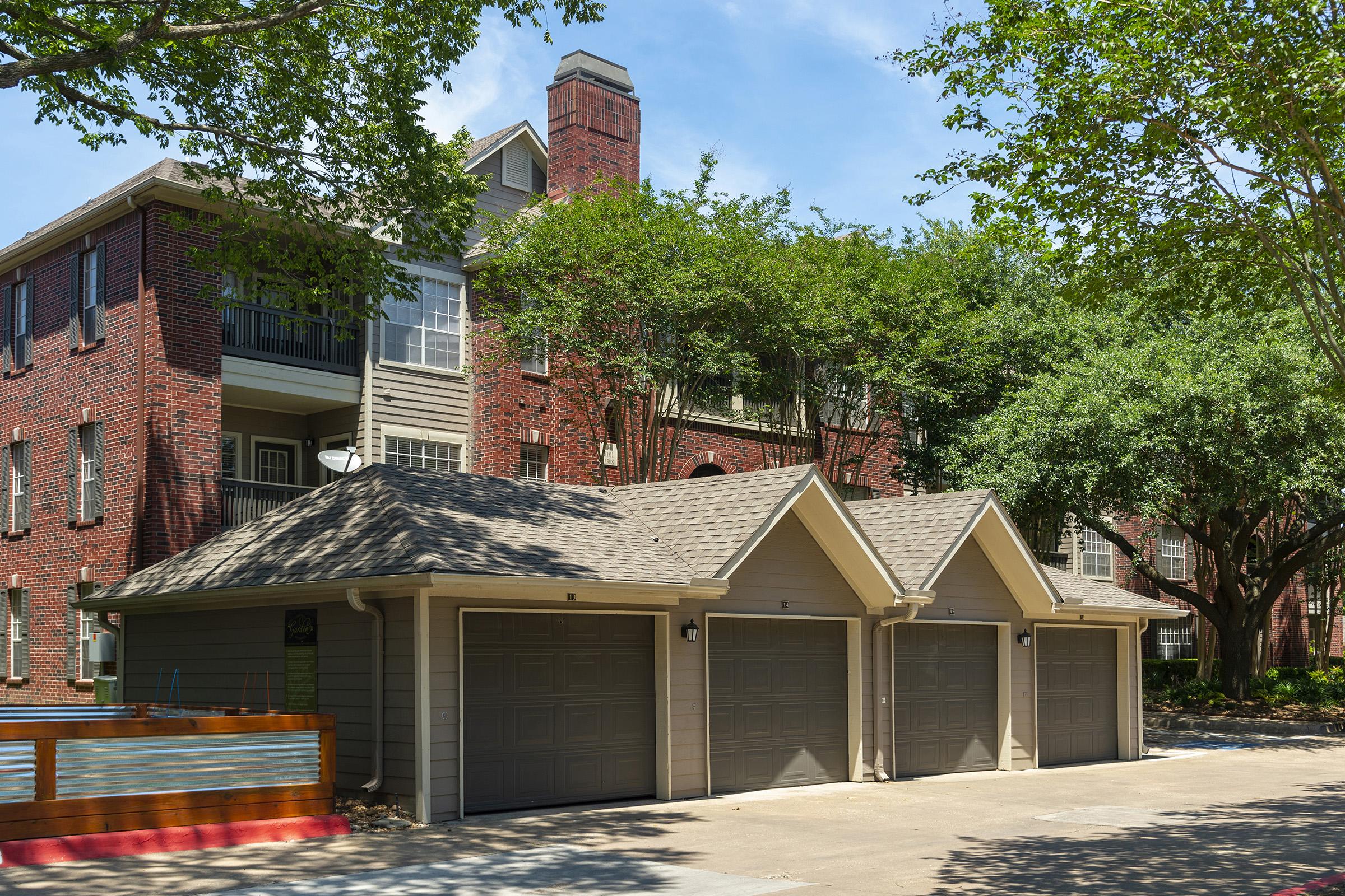 Two garage doors in front of a multi-story brick apartment building surrounded by lush green trees. The structure features a modern design with balconies, and the driveway is paved. Bright sunlight casts shadows on the ground, creating a vibrant outdoor atmosphere.