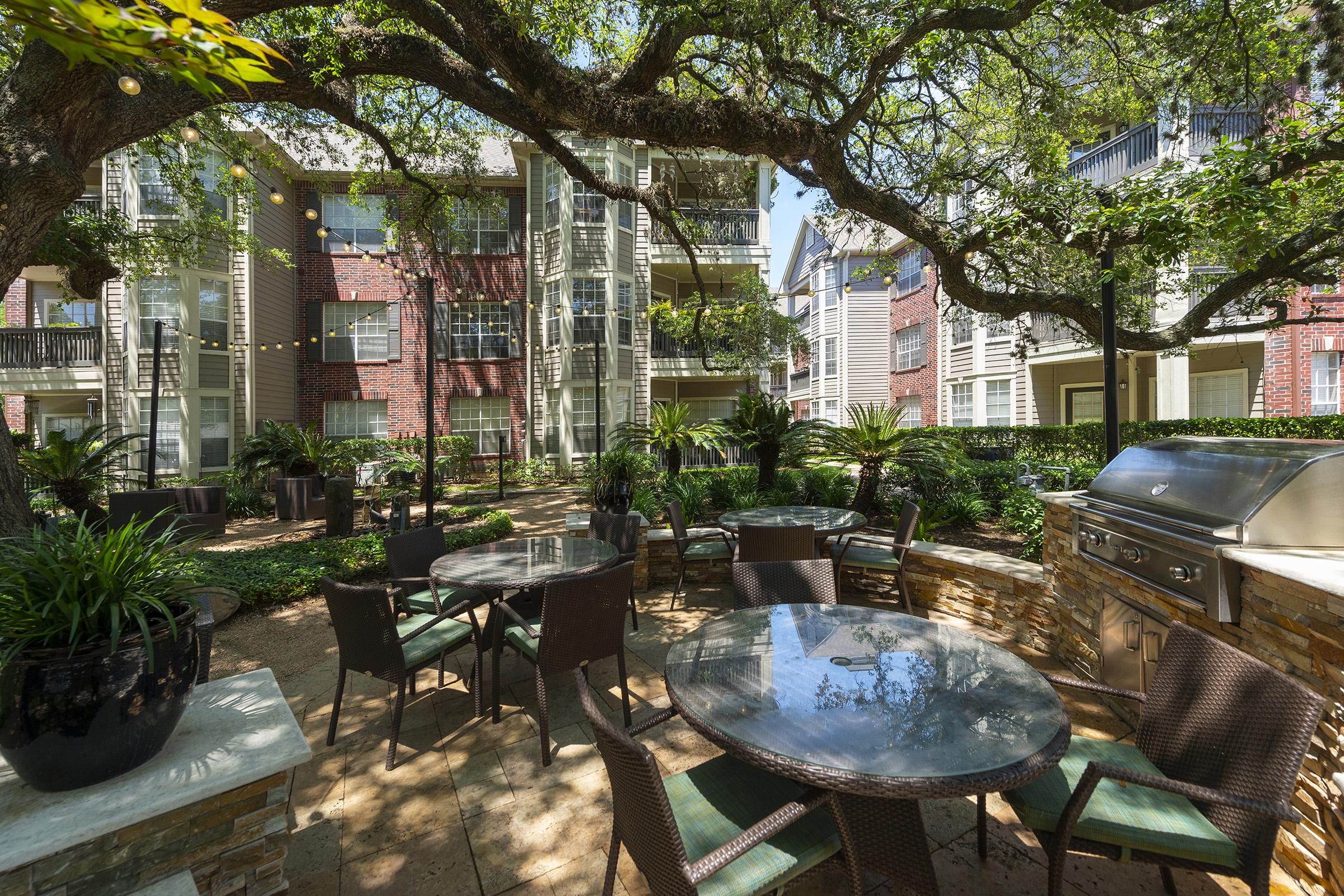 A shaded outdoor space featuring multiple round tables with chairs, surrounded by greenery and shrubs. In the background, brick apartment buildings are visible, along with a barbecue grill. The area is well-lit and invites relaxation, ideal for gatherings or outdoor dining.