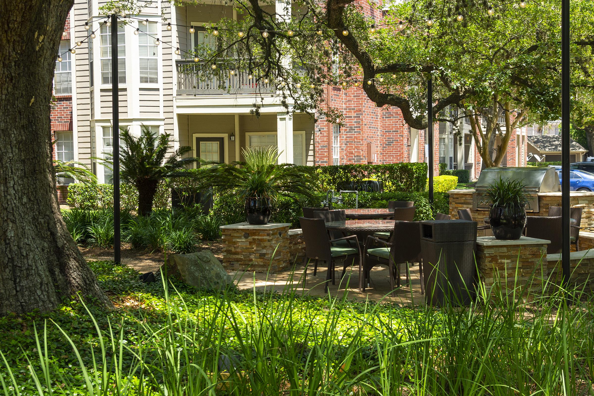 A landscaped outdoor space featuring green grass, shrubs, and small palm trees. In the foreground, there is a dining area with a table and chairs surrounded by stone planters. In the background, apartment buildings are visible, partially shaded by large trees. Decorative string lights are hung above the seating area.