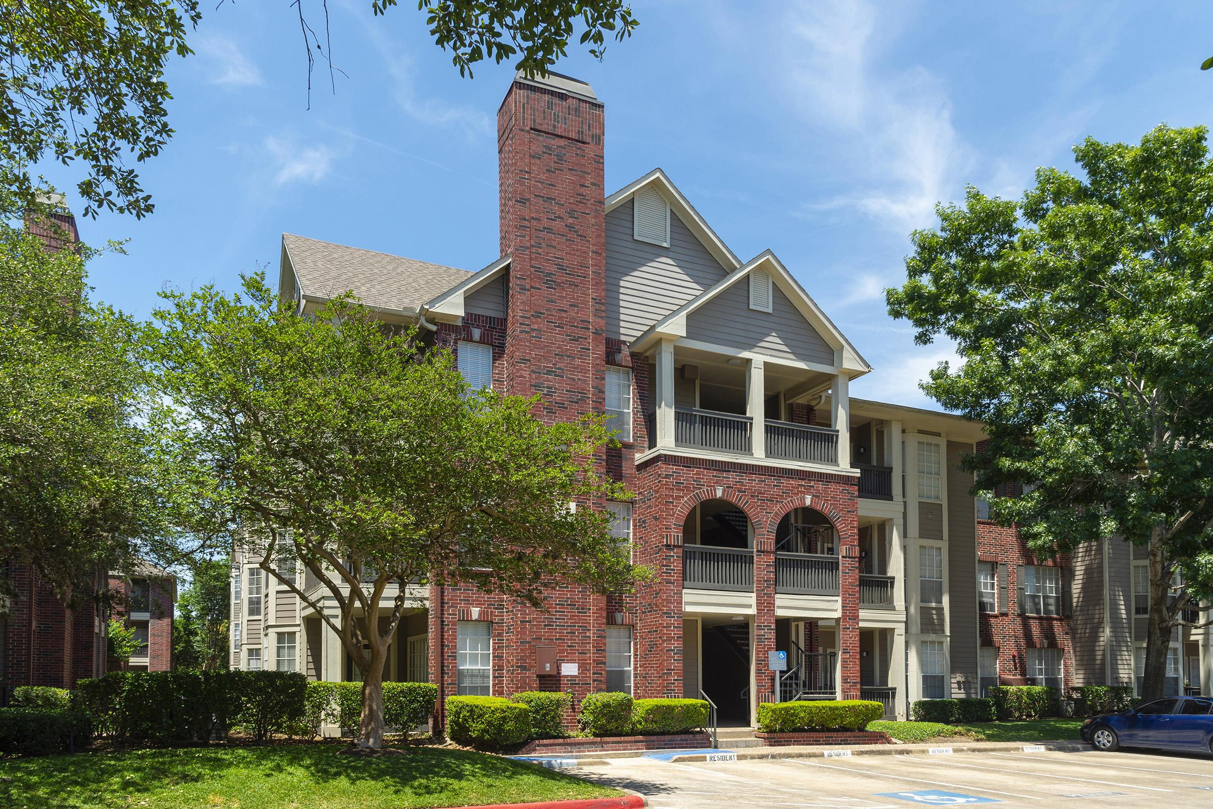 A three-story brick apartment building featuring a tall chimney, arched balconies, and landscaped greenery. The setting is sunny with a clear blue sky, and parking spaces are visible in the foreground.