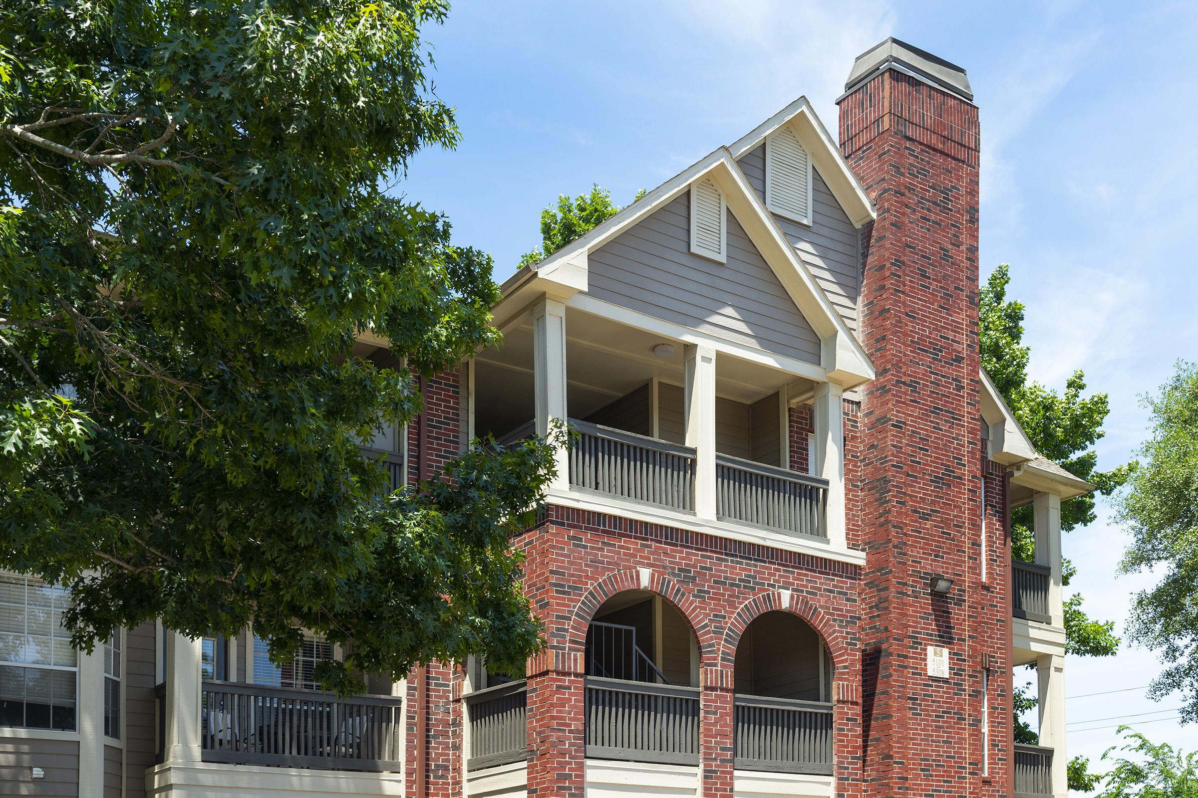 A view of a multi-story brick building with large balconies, surrounded by trees and green foliage. The architecture features a mix of modern and traditional styles, with a clear blue sky in the background, creating a bright and inviting atmosphere.
