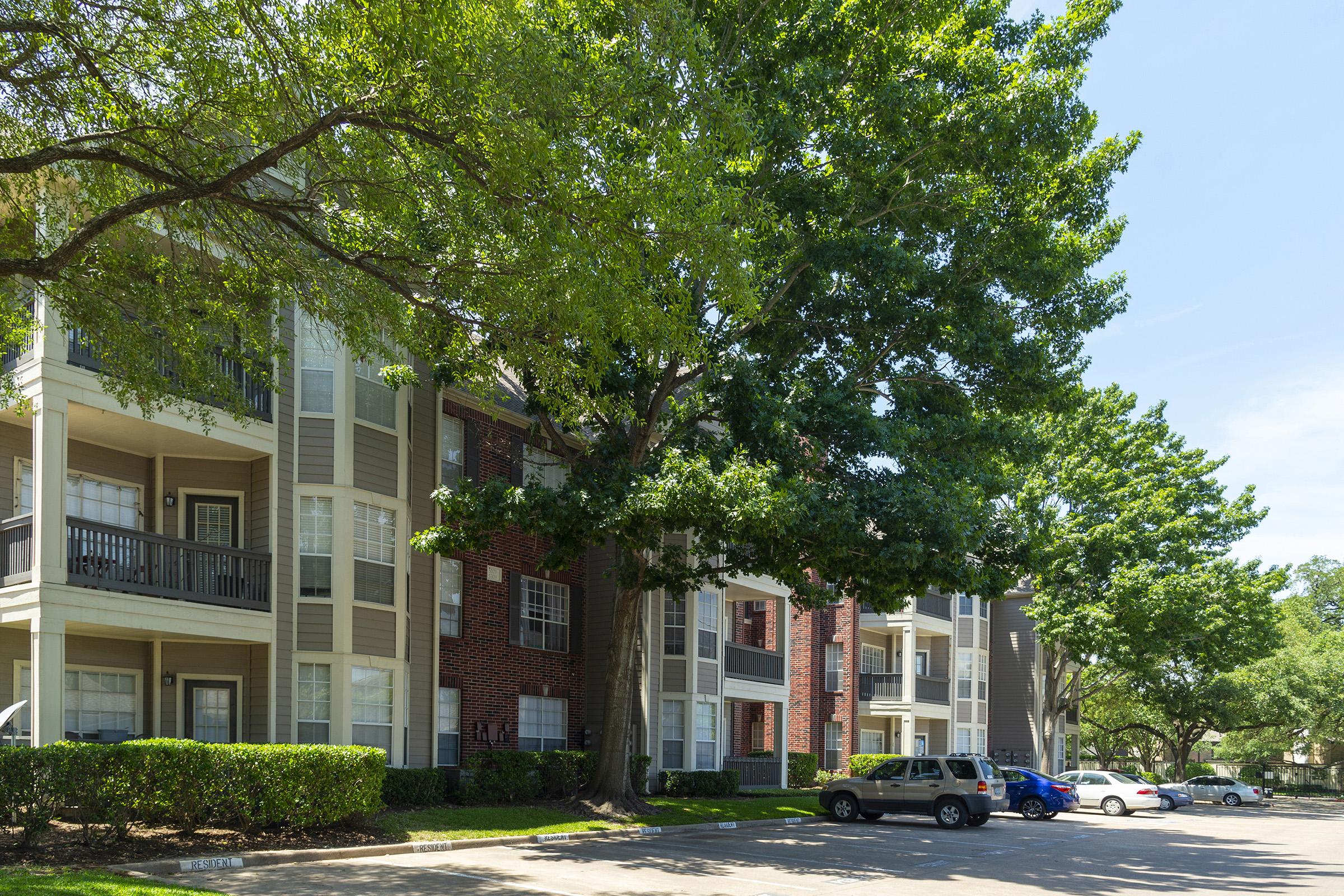 A residential apartment building featuring a mix of brick and siding exteriors, surrounded by lush green trees and shrubs. The scene includes parked cars in the foreground and bright blue sky overhead, conveying a calm and inviting atmosphere.