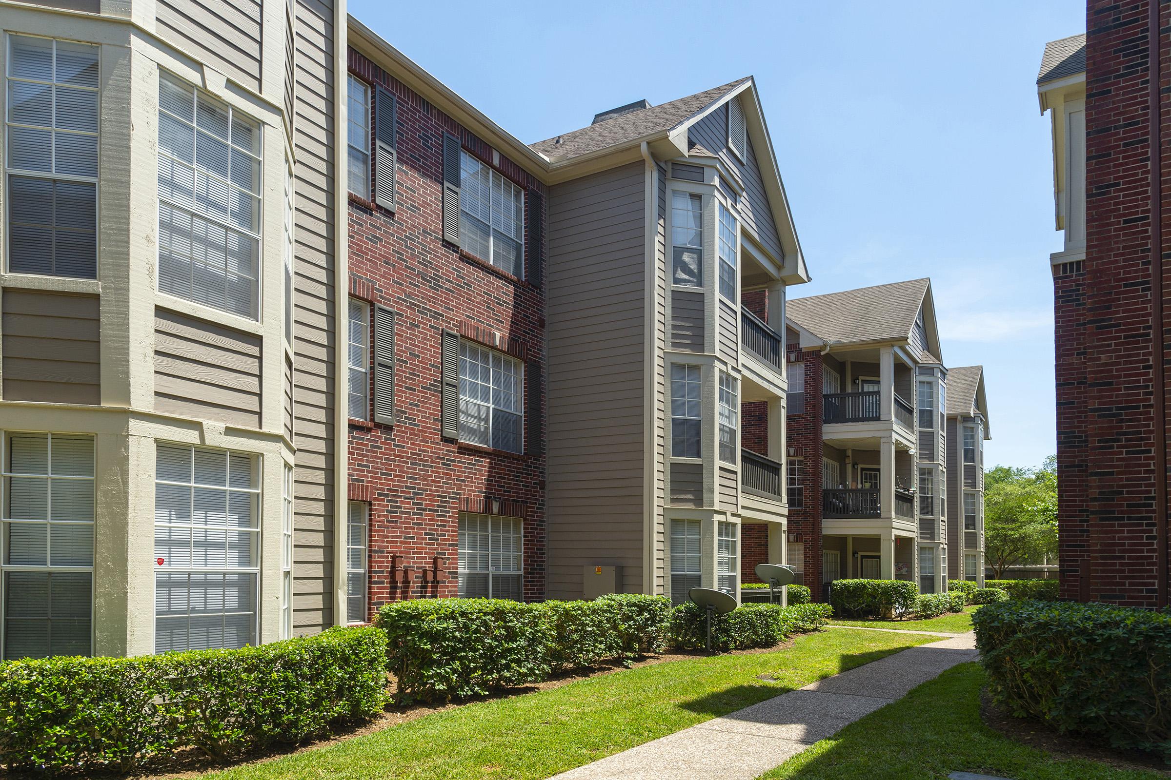A well-maintained apartment complex featuring two adjacent buildings with a mix of brick and siding exteriors. Lush green hedges line the pathway leading to the entrance, with balconies visible on the upper floors. The scene is set on a sunny day with clear blue skies.