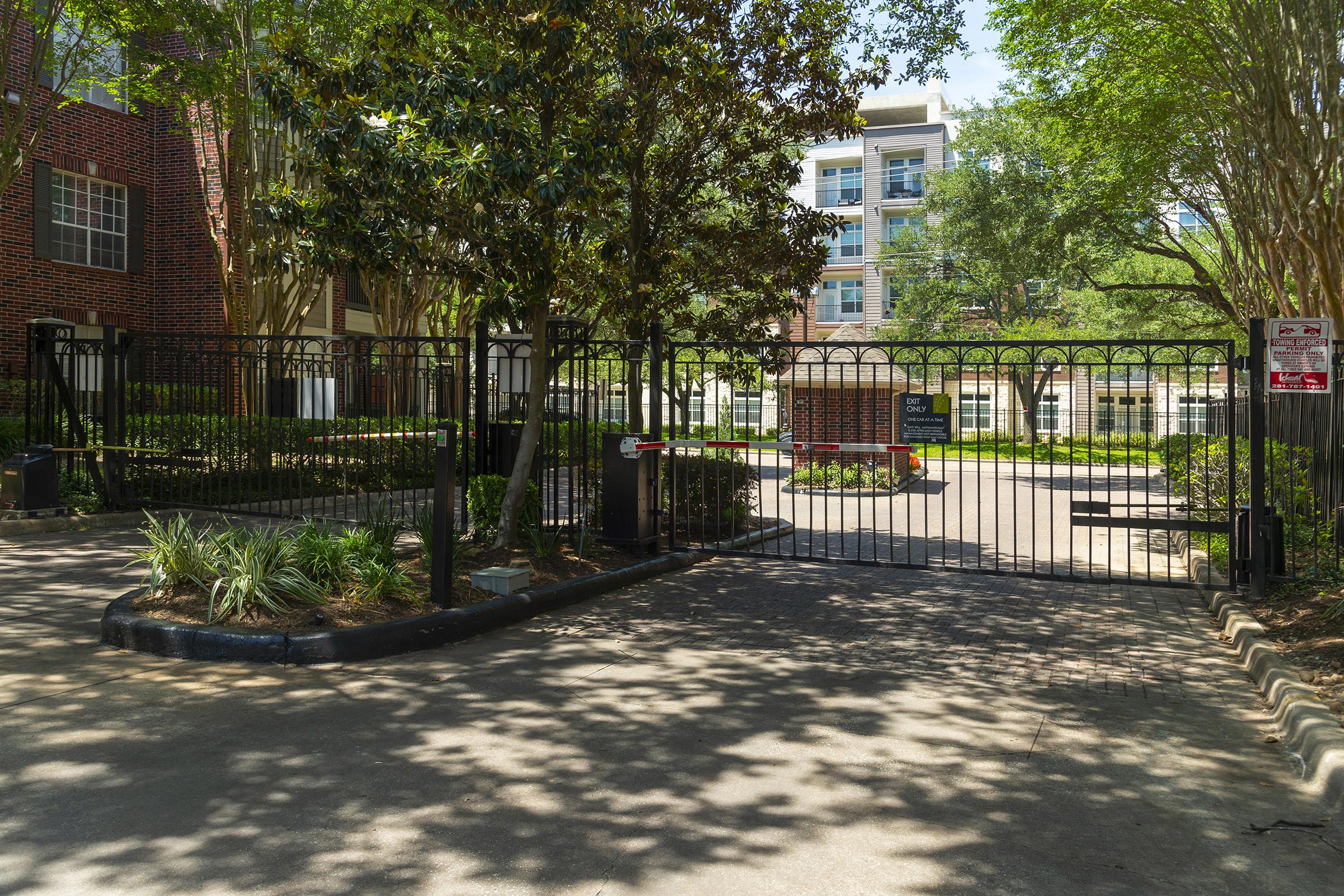 Gated entrance to a residential complex, featuring a black iron gate surrounded by landscaped greenery. Sunlight casts shadows on the paved driveway, with a glimpse of modern buildings visible in the background, suggesting an urban setting.