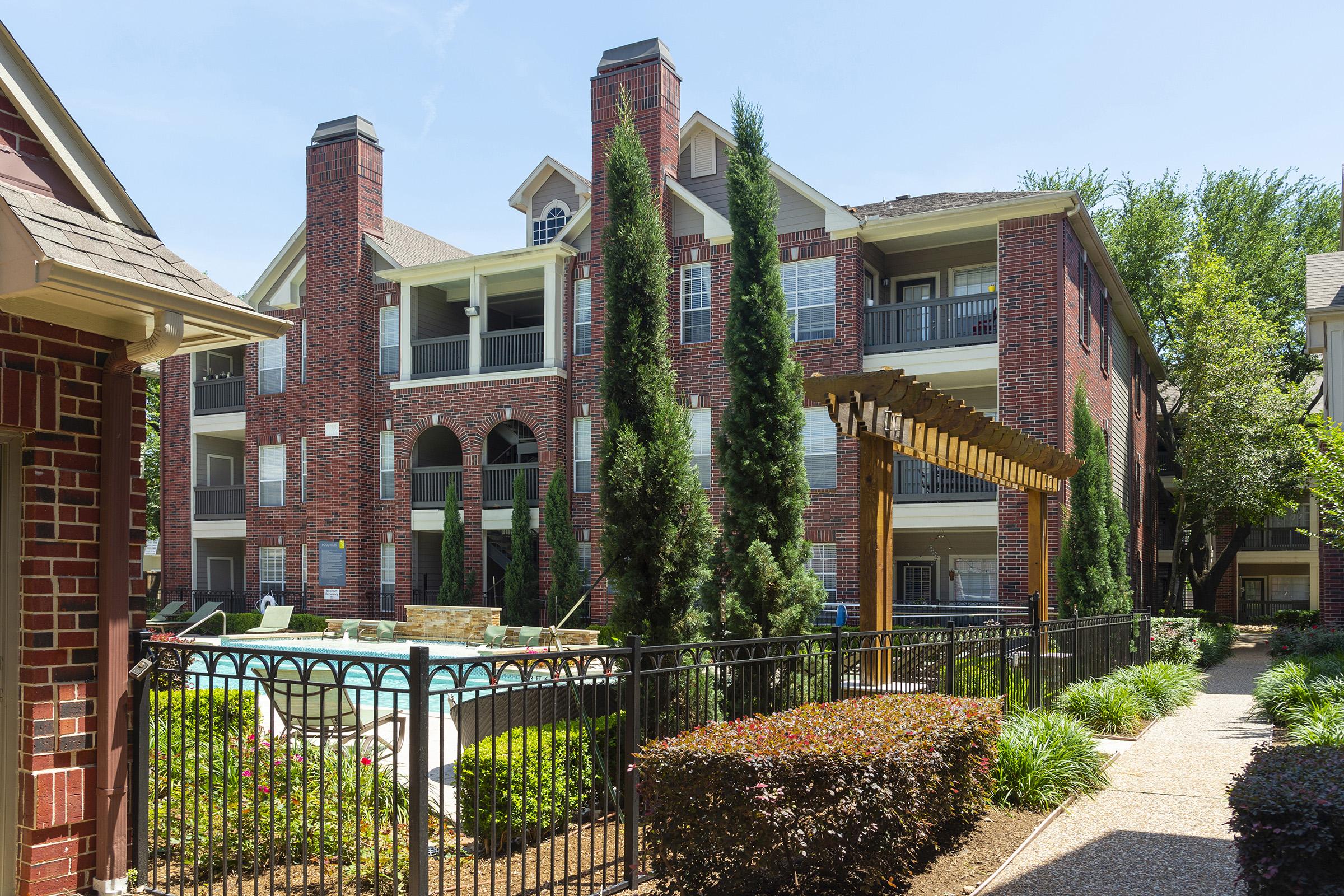 View of a multi-story brick apartment building with balconies, surrounded by tall evergreen trees and a well-maintained landscape. In the foreground, a decorative wooden pergola frames a glimpse of a swimming pool area. Clear blue sky overhead adds to the bright, inviting atmosphere.