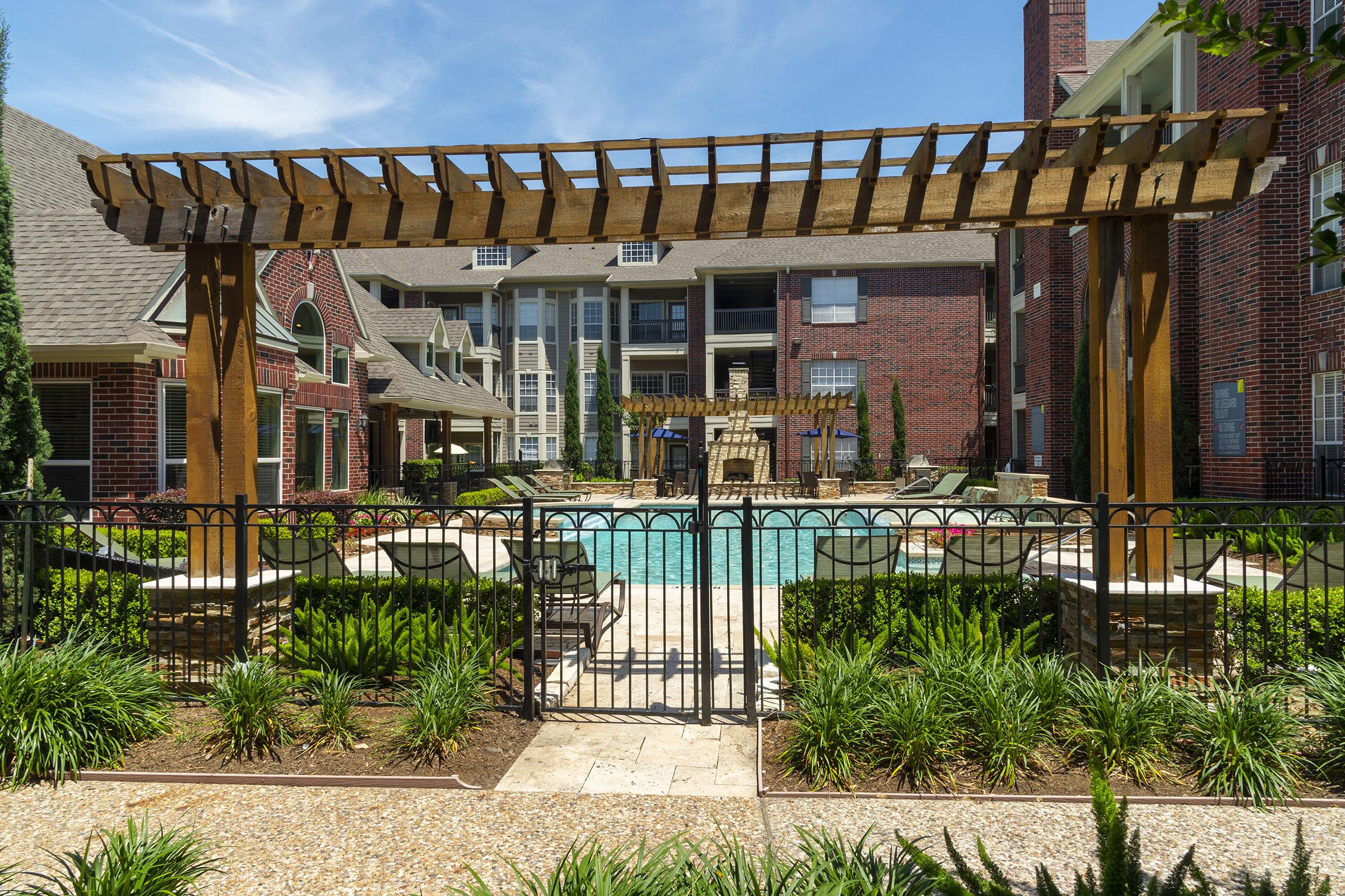 A sunny courtyard featuring a swimming pool surrounded by lounge chairs and a stone patio. A wooden trellis arches over a seating area next to the pool, with well-maintained greenery and shrubs framing the scene. Brick buildings in the background complete the inviting residential atmosphere.