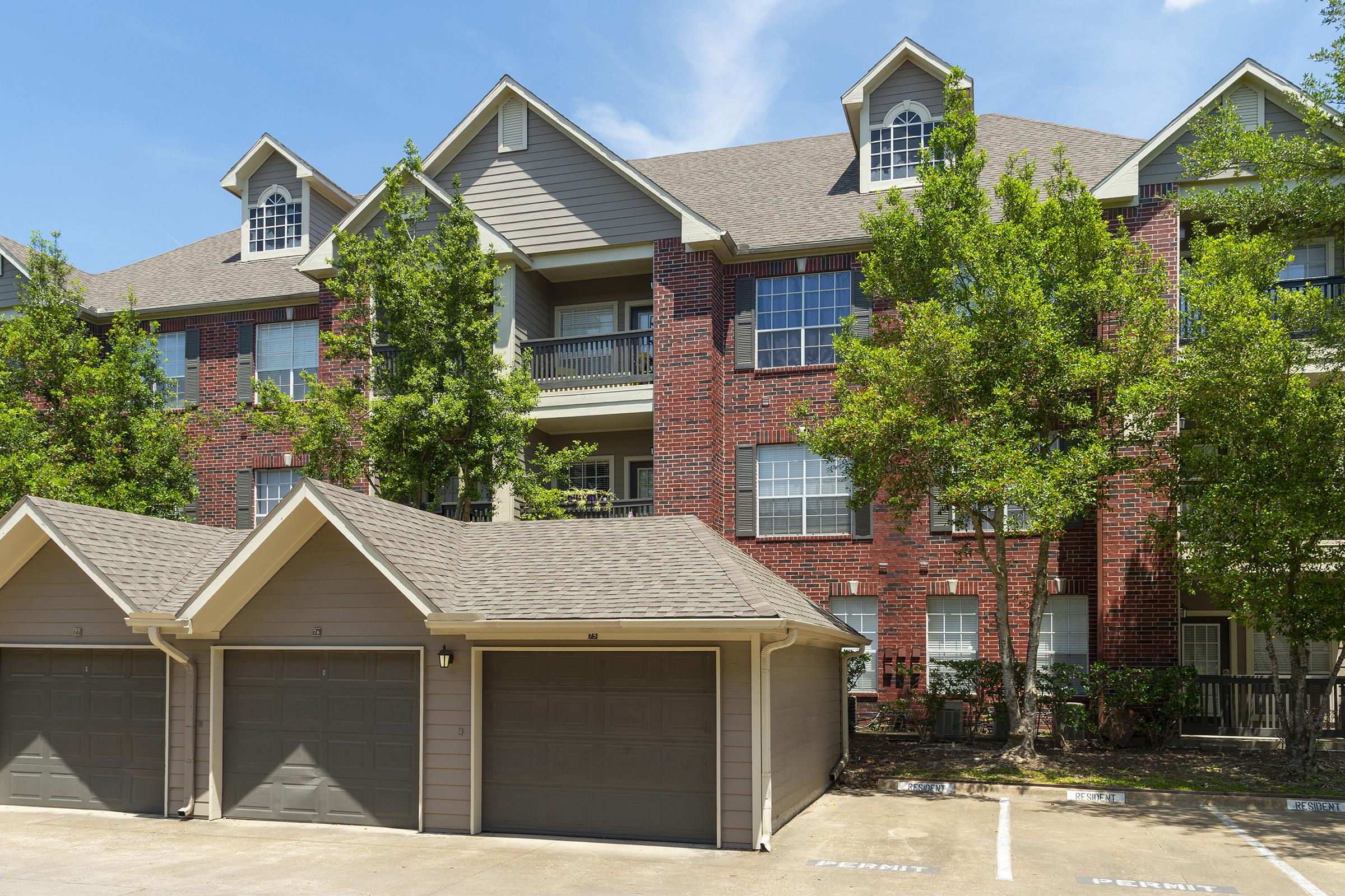 A multi-story apartment building with a mix of brick and siding exterior, featuring large windows and balconies. In the foreground, there are three garage doors, and the surrounding area includes well-maintained trees and landscaping under a clear blue sky.