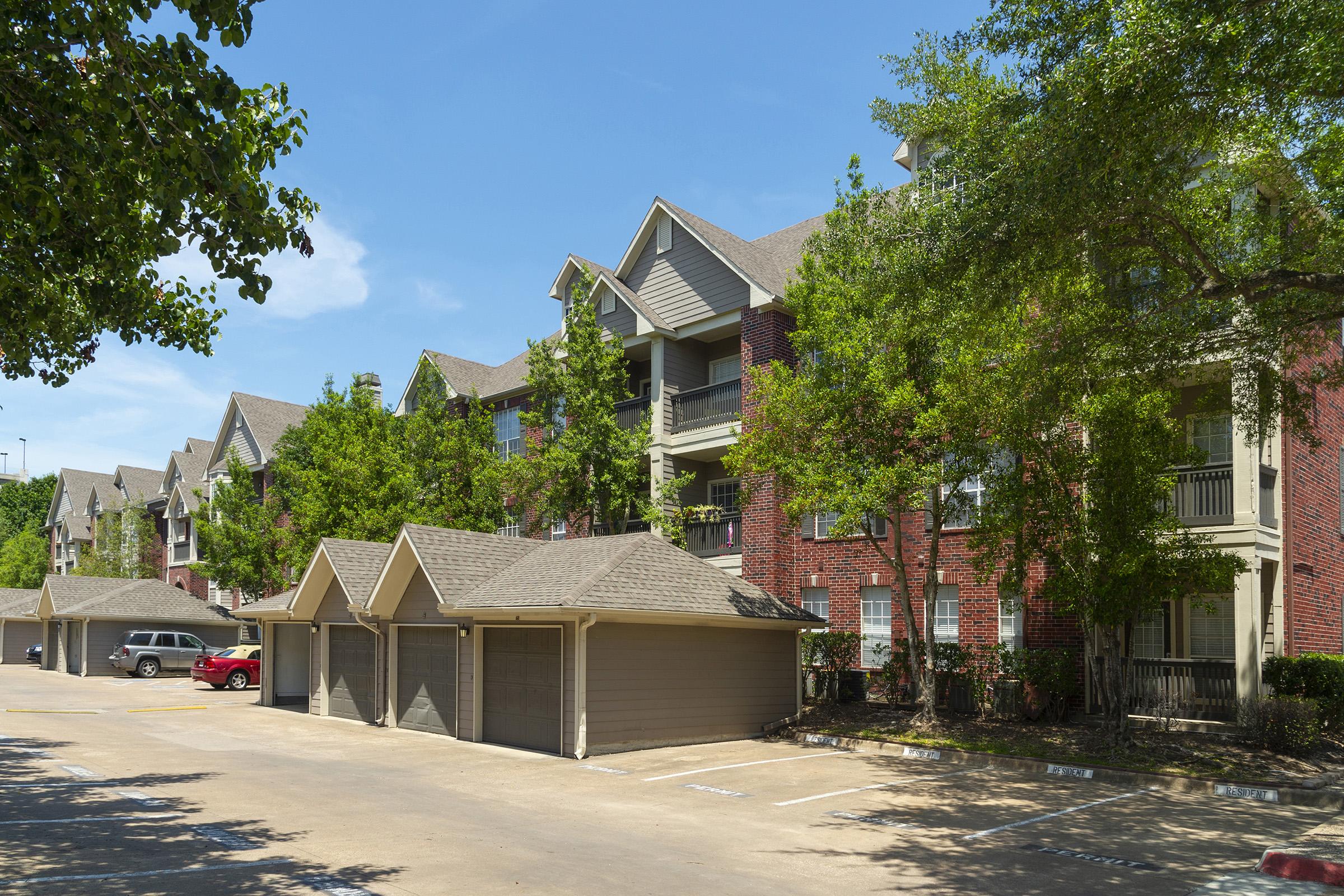 A row of three-story apartment buildings with a mix of brick and siding exteriors. The buildings are surrounded by mature trees and shrubs. In the foreground, there are parking spaces with two parked cars and several garage doors. The sky is clear and blue, indicating a sunny day.