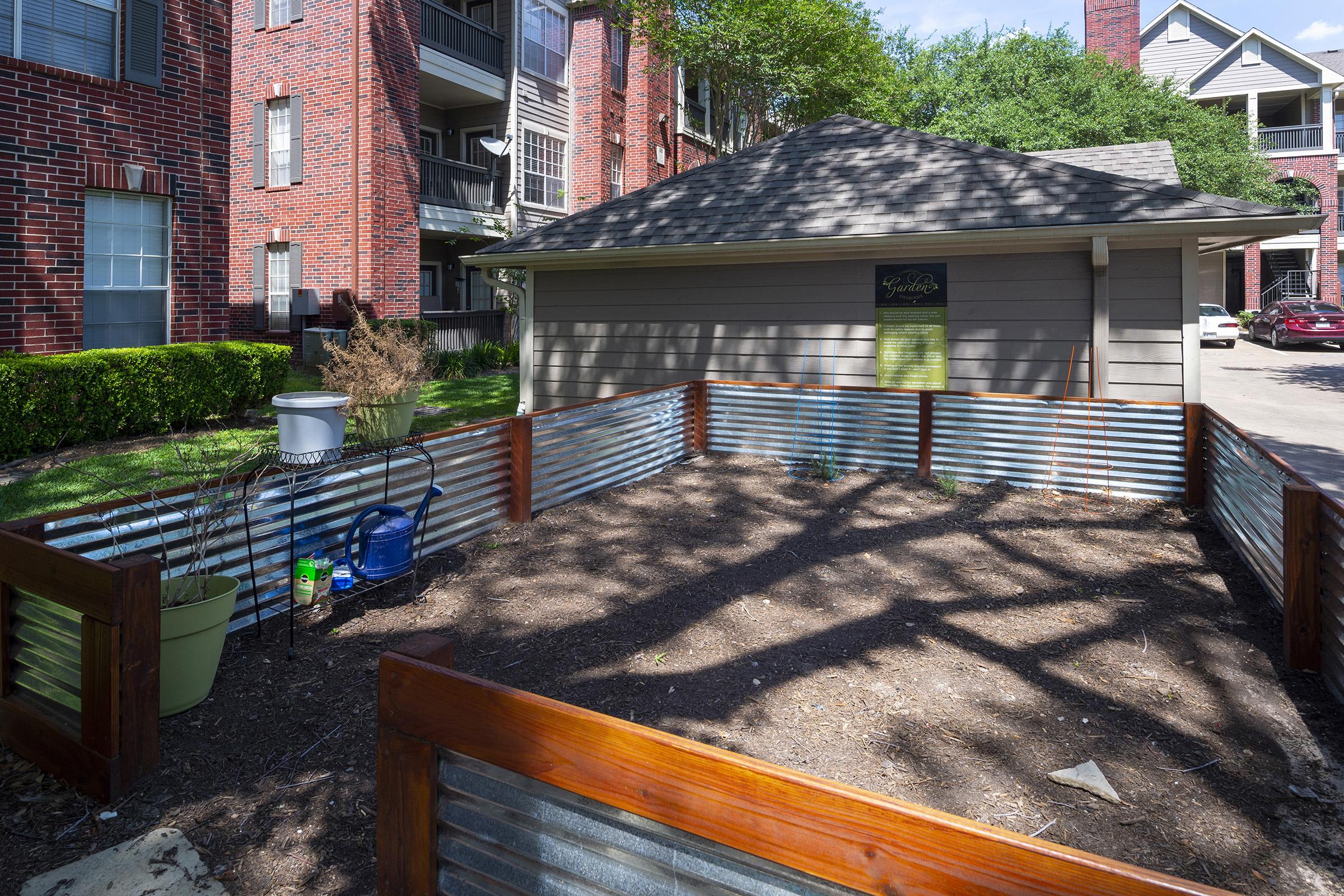 Community garden space featuring raised garden beds made of corrugated metal and wooden borders. The area is surrounded by red-brick apartment buildings and has a shed with a sign. There are gardening tools and a white bucket placed nearby, with trees casting shadows over the soil.