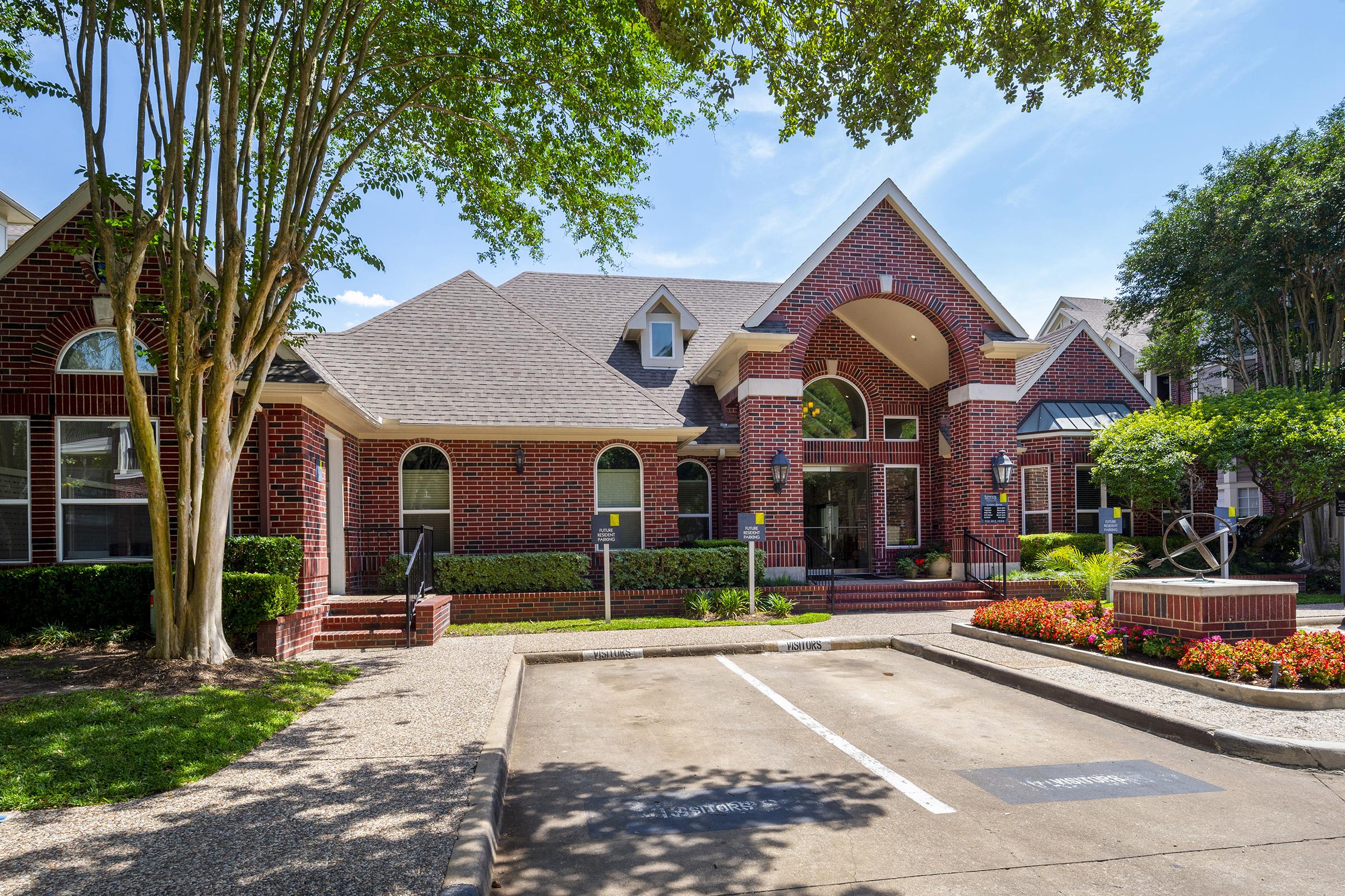 A red brick building with a triangular roof and large windows, surrounded by lush trees and colorful flower beds. The entrance features a decorative sculpture and a spacious parking area with visible parking signs. The scene is bathed in bright sunlight, giving a welcoming atmosphere.