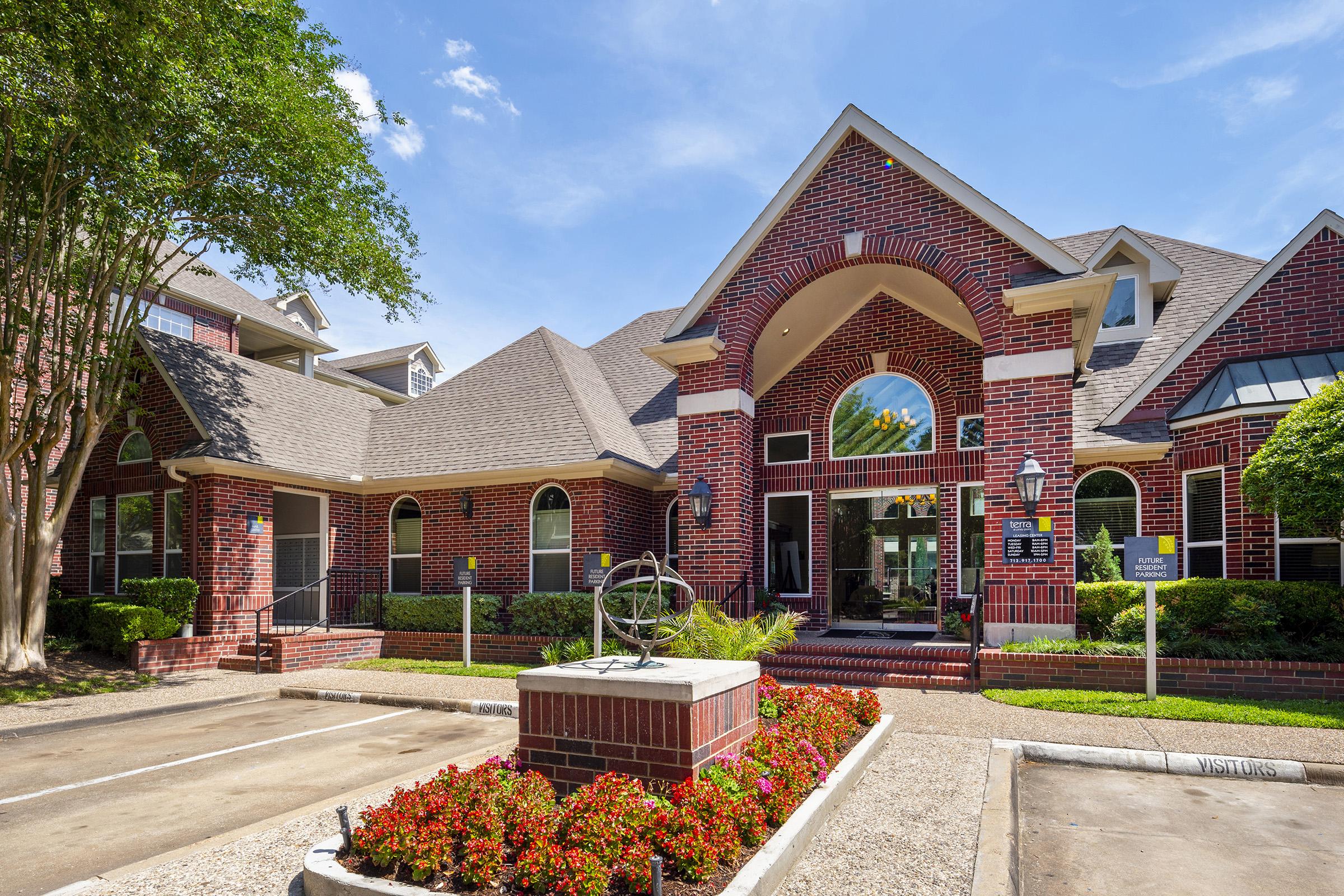 A brick building featuring large arched windows and a decorative entrance. In front, there is a landscaped area with colorful flowers and a sundial sculpture. The sky is clear with a few clouds, and the surroundings include green trees and a parking area with visitor signage.