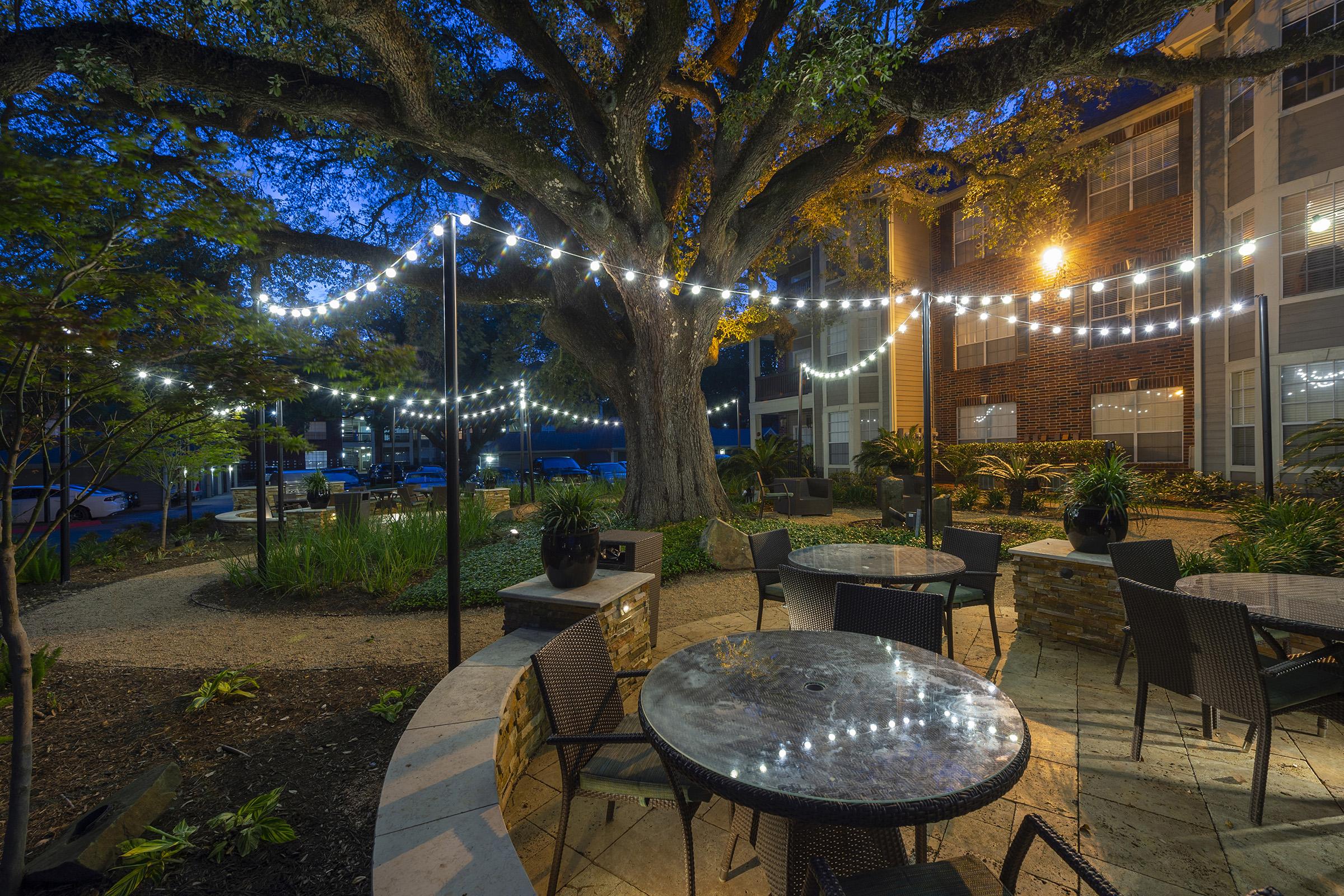 A cozy outdoor space at dusk featuring a large tree, string lights, and round tables with chairs. The area is surrounded by lush greenery and has a warm, inviting atmosphere, perfect for socializing or relaxation.