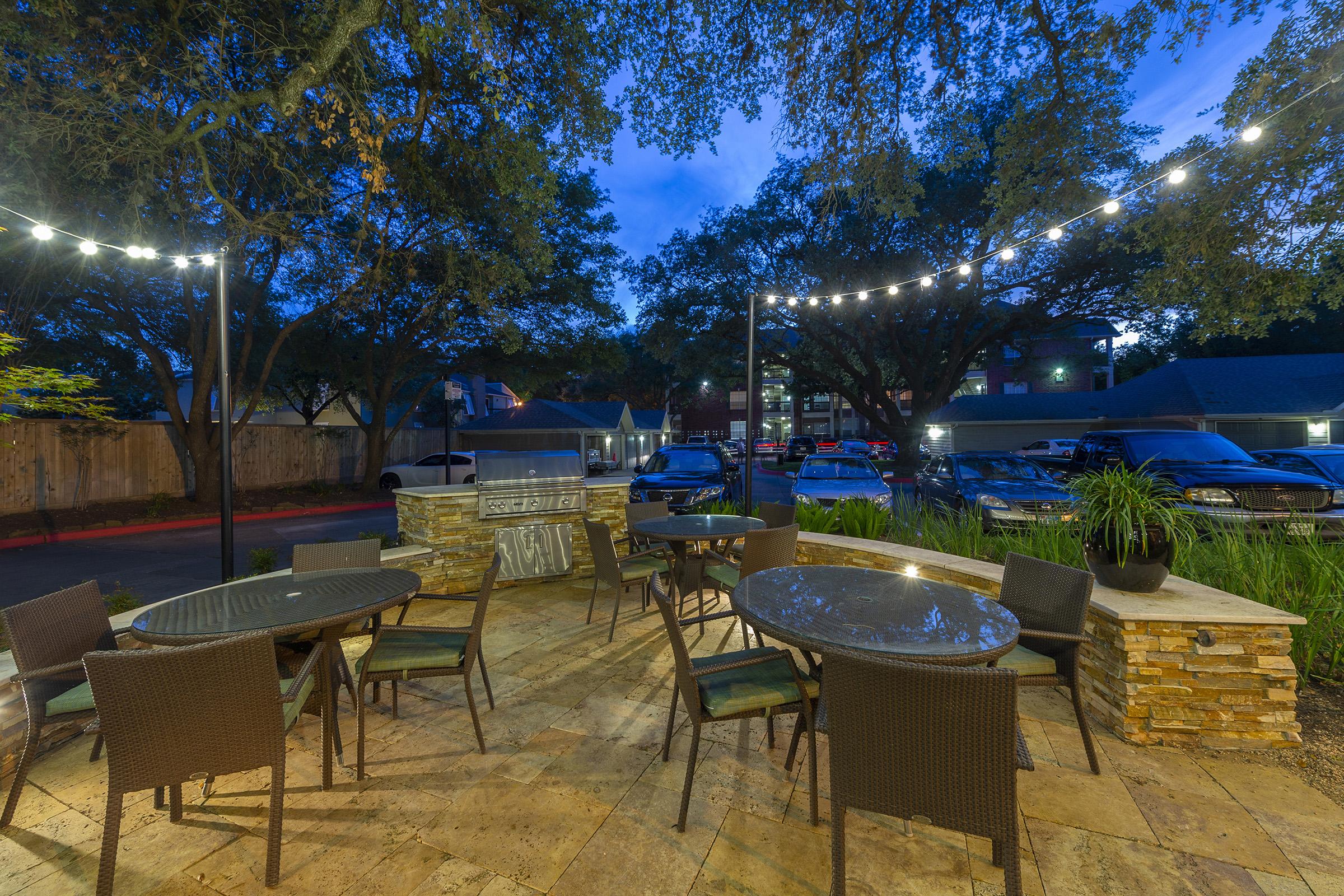 Outdoor seating area with stone tables and chairs, surrounded by greenery. Warm string lights are strung above, creating a cozy ambiance during twilight. In the background, parked cars and buildings are visible, adding to the inviting atmosphere.