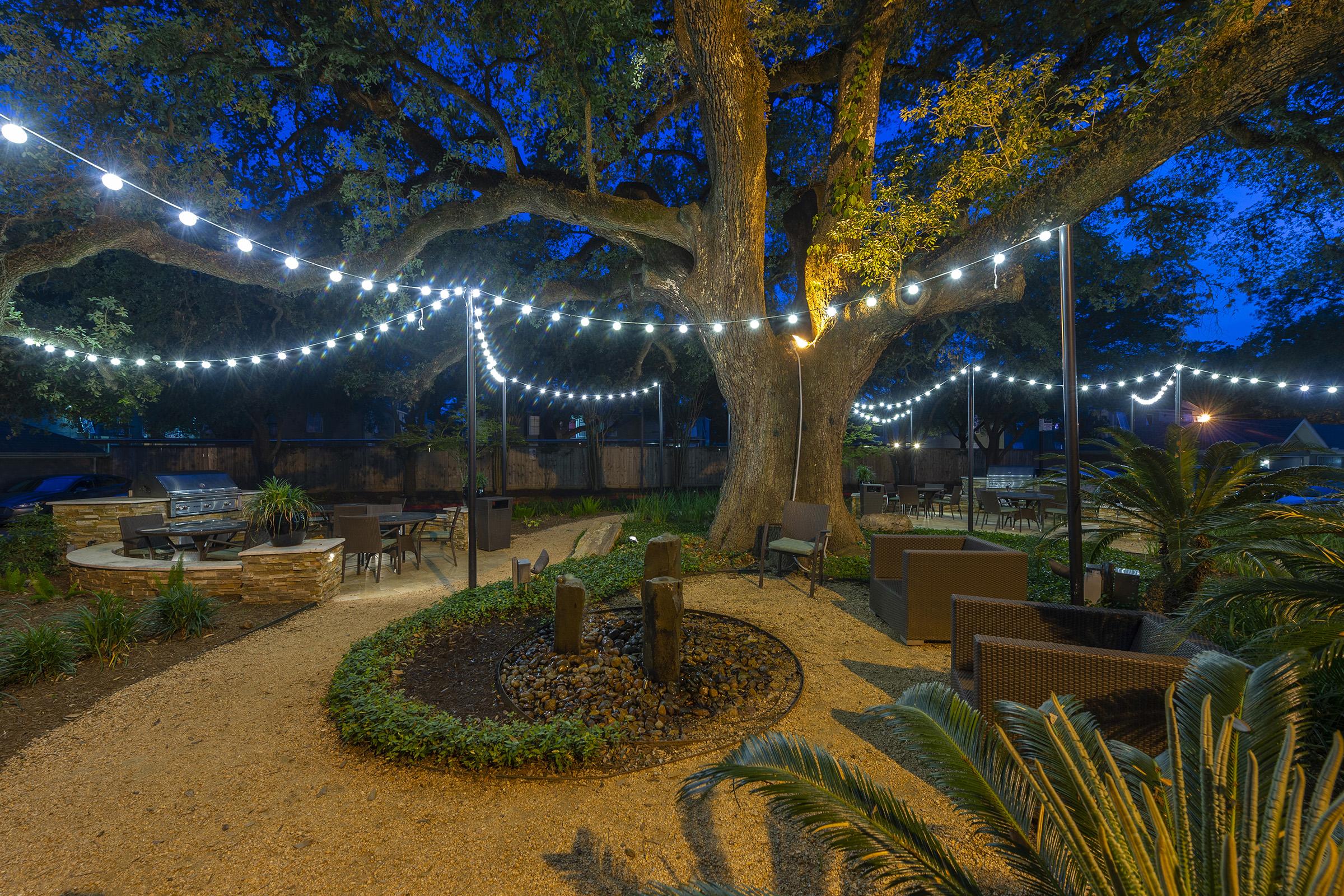 Outdoor garden area at night with a large tree adorned with string lights. Gravel pathways wind through greenery and seating arrangements, including lounge chairs and a central fire pit. The ambiance is tranquil, illuminated by warm light, creating a cozy atmosphere for relaxation.
