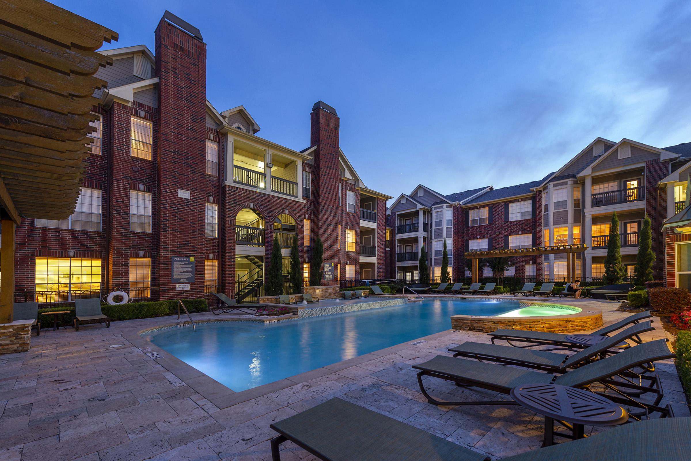 A tranquil evening view of a luxury apartment complex featuring a sparkling swimming pool surrounded by lounge chairs. The buildings are constructed with red brick, and lush greenery decorates the area. Soft lighting from the windows creates a warm ambiance, highlighting the inviting atmosphere of the community.