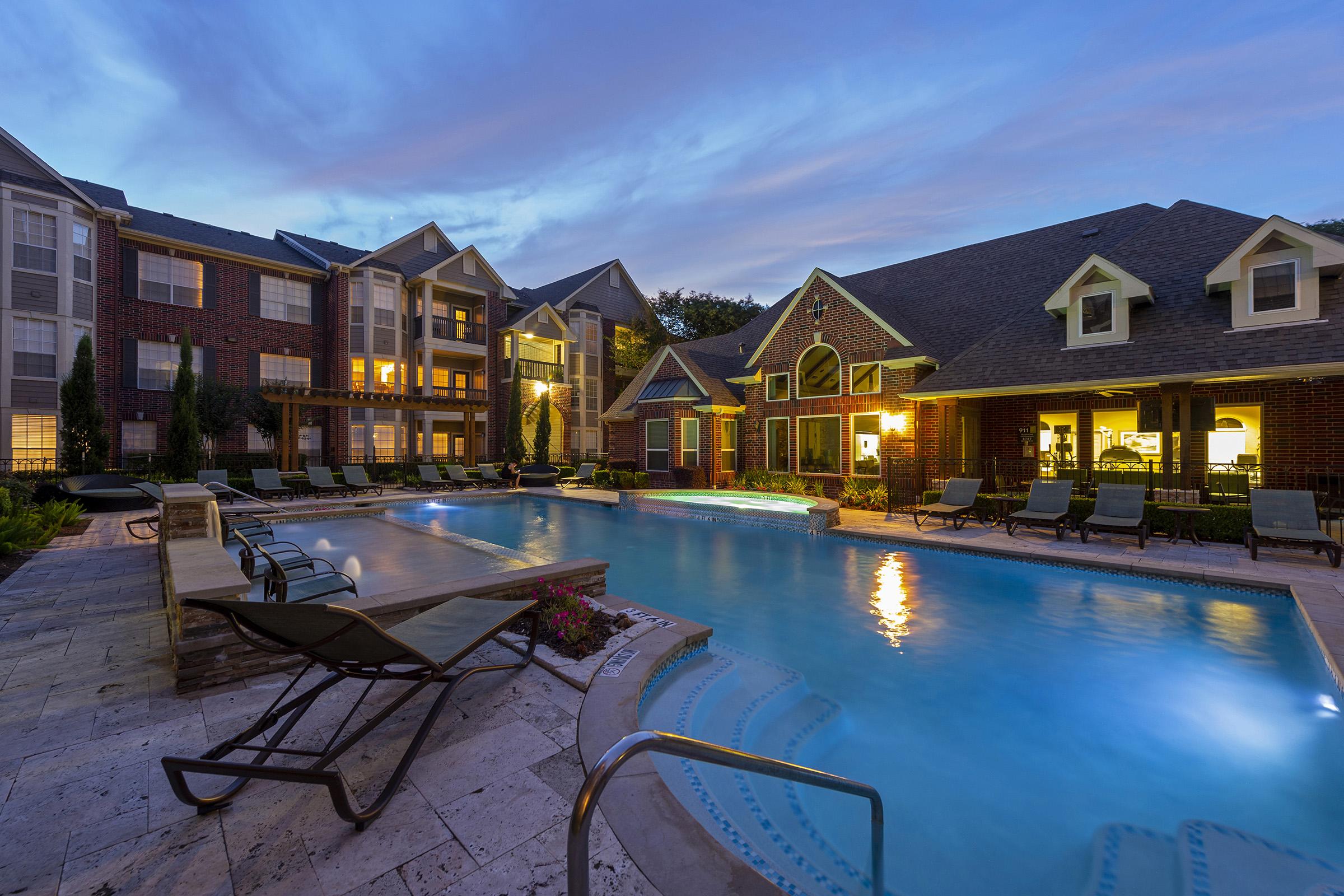 A tranquil evening scene featuring a well-lit outdoor swimming pool surrounded by lounge chairs, with a brick apartment complex in the background. The pool reflects the soft glow of the lights and the sky is painted in hues of blue and purple, creating a serene atmosphere for relaxation.