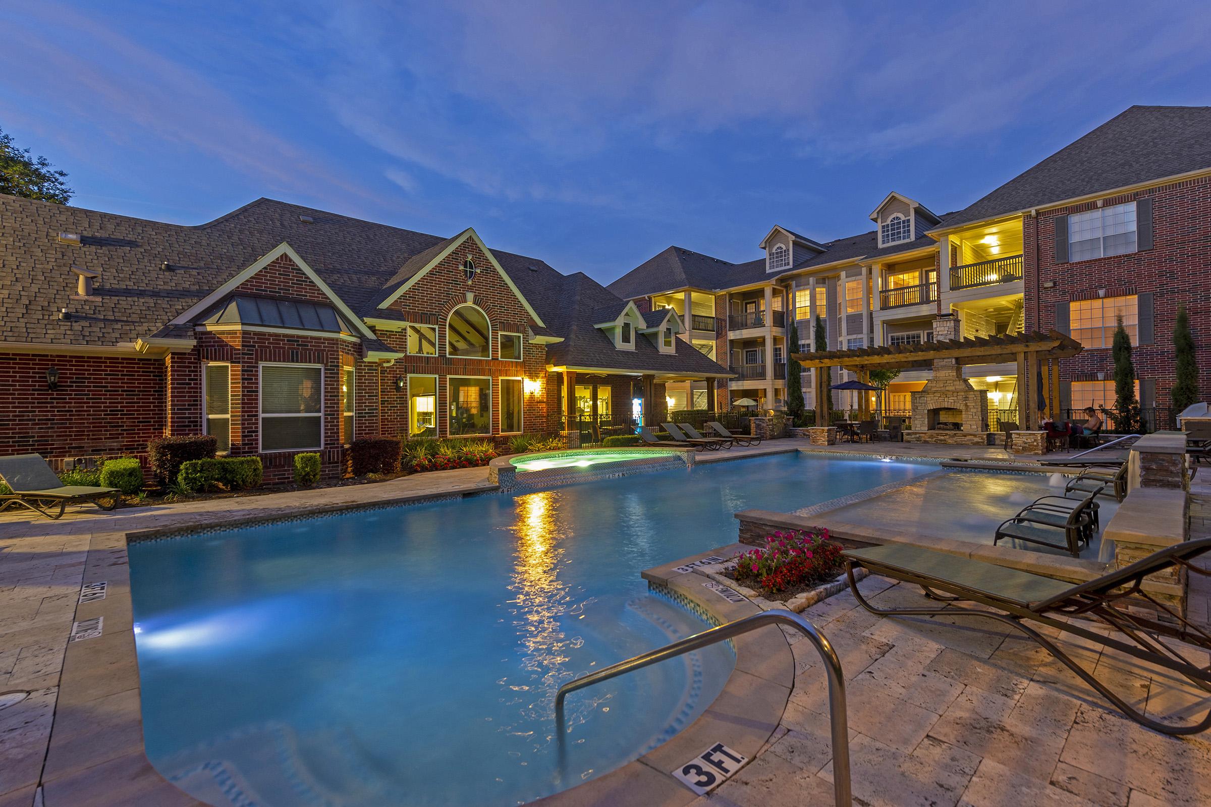 A beautifully lit pool area at dusk, featuring a sparkling blue pool with lounge chairs surrounding it. In the background, elegant brick apartment buildings are visible, with warmly lit windows and a cozy outdoor seating area. The scene conveys a relaxed and inviting atmosphere.