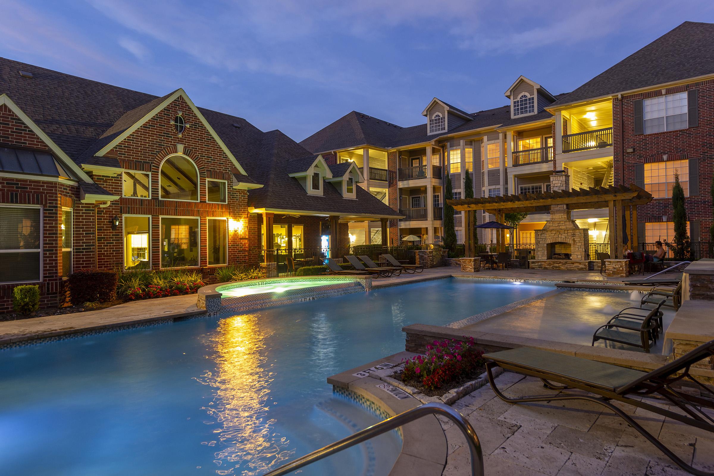 A beautifully designed apartment complex featuring a luxurious pool area at dusk. The pool is illuminated with soft lights, surrounded by lounge chairs and vibrant landscaping. The buildings showcase elegant architecture with balconies and warm lighting, creating a welcoming atmosphere.