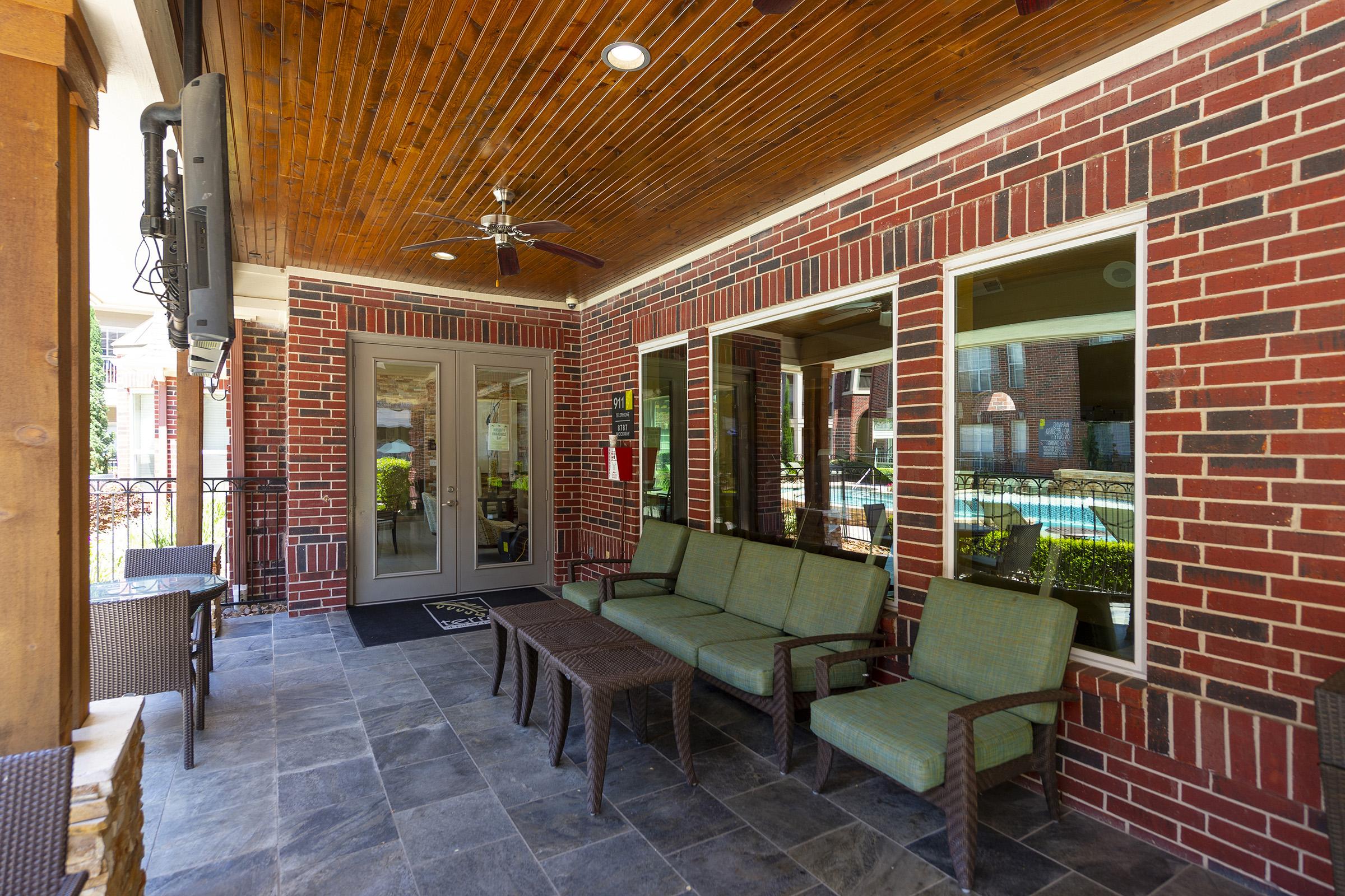 A cozy outdoor lounge area with a stone floor, featuring a wicker coffee table and green cushioned seating. The walls are made of red brick, complemented by large windows that offer views of the outside. A ceiling fan hangs above, and a television is mounted on the wall.
