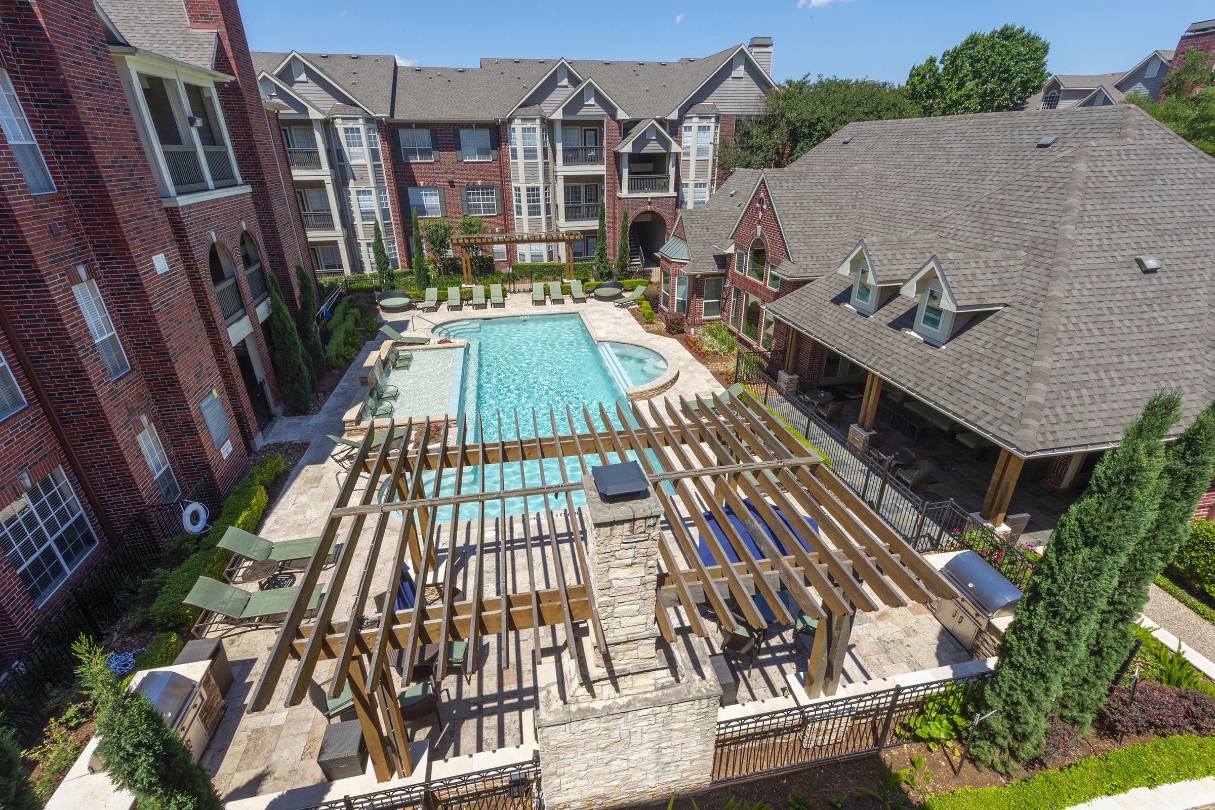 Aerial view of a residential apartment complex featuring a large swimming pool surrounded by lounge chairs and a pergola. Nearby, there are landscaped areas and buildings with brick facades. The scene is bright and sunny, showcasing a relaxing outdoor community space.