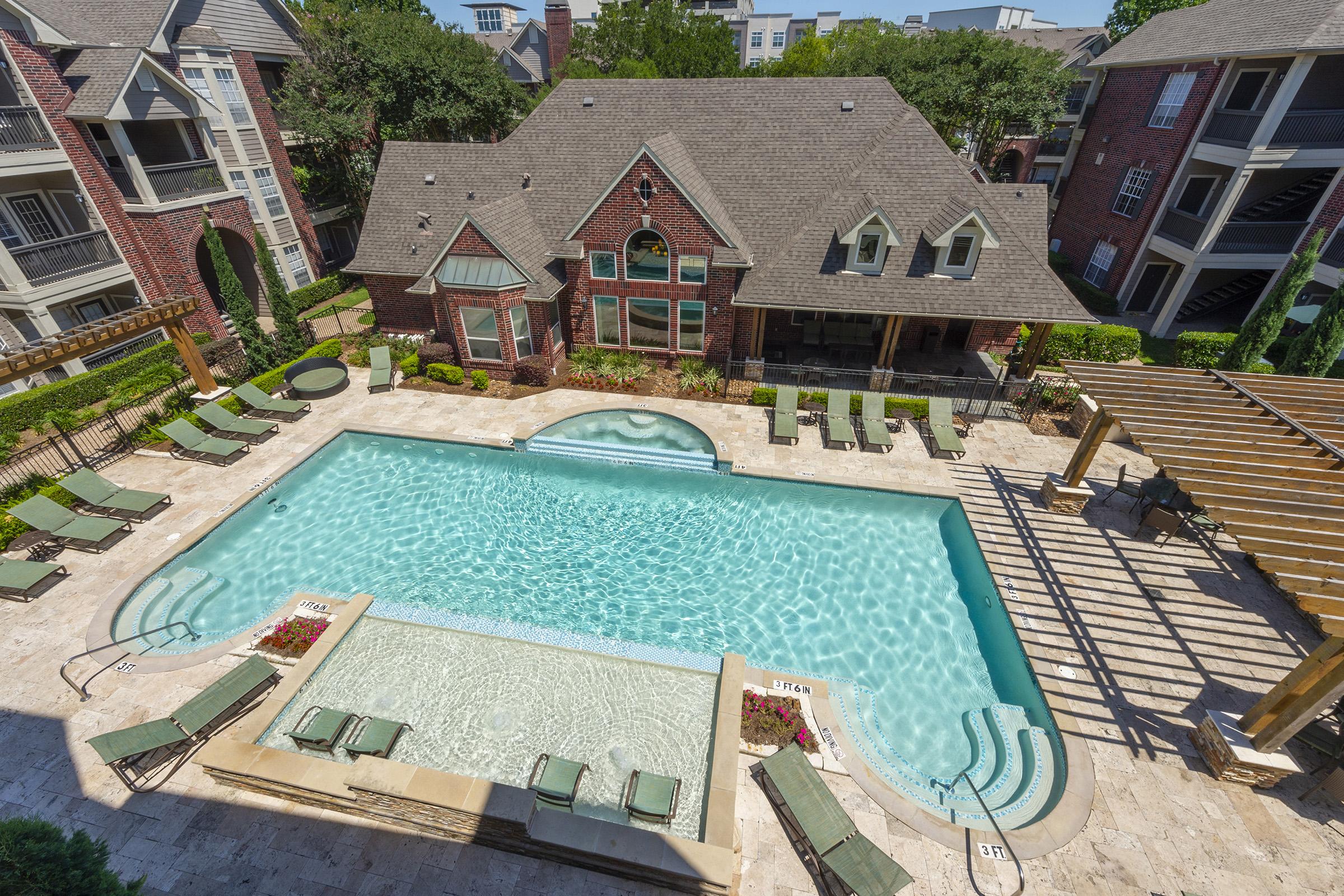 Aerial view of a swimming pool surrounded by lounge chairs in a residential community. A spacious brick clubhouse with large windows is visible in the background, along with shaded pergolas near the pool area. Lush greenery and landscaping enhance the inviting atmosphere.