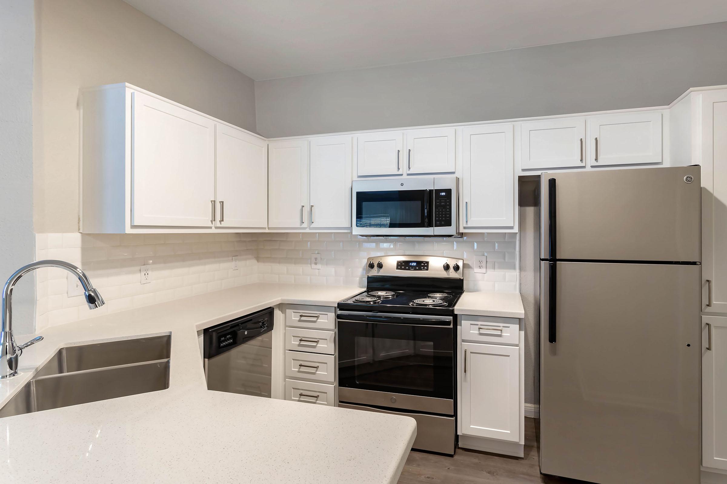 Modern kitchen featuring white cabinetry, stainless steel appliances including a microwave, oven, and refrigerator. A sleek countertop and a stainless steel sink complement the design, with a neutral wall color adding to the contemporary feel. Bright and inviting, perfect for cooking and entertaining.