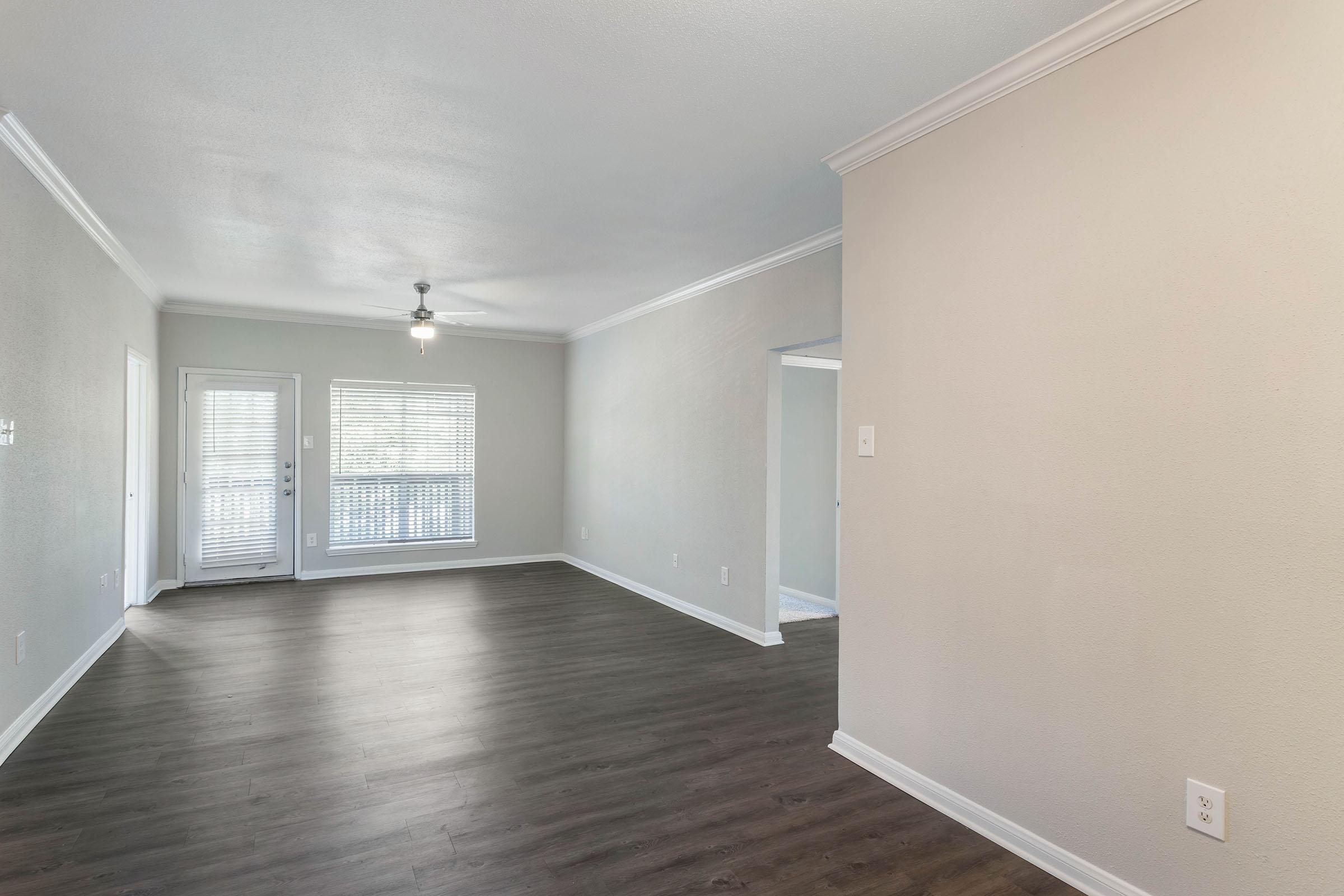 Spacious, empty living area featuring light-colored walls, a ceiling fan, and large windows allowing natural light. The room has new laminate flooring and leads to a balcony through sliding glass doors. A doorway to another room is visible in the background.