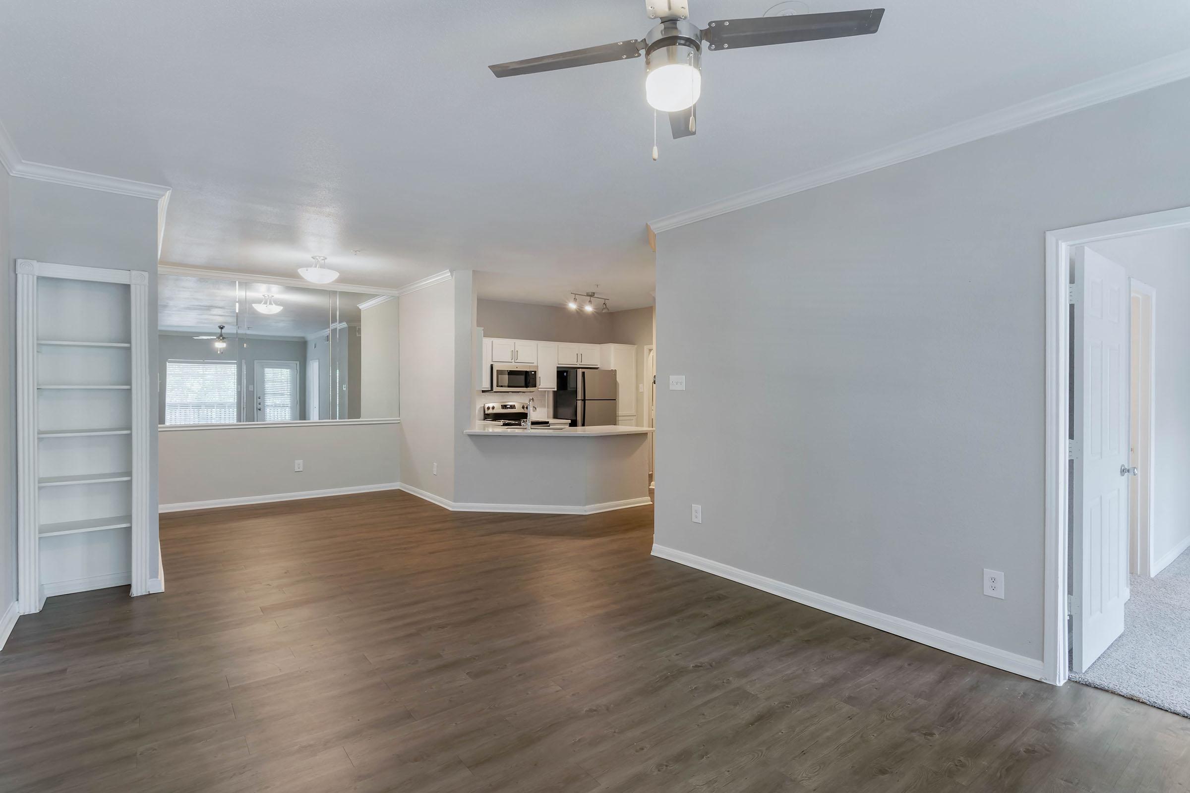 Spacious living area with wood-style flooring, ceiling fan, and neutral-colored walls. Features an open layout leading to a kitchen with modern appliances and a breakfast bar. Natural light enters through large windows, and a built-in bookshelf is visible in the corner. Perfect for comfortable living or entertaining.