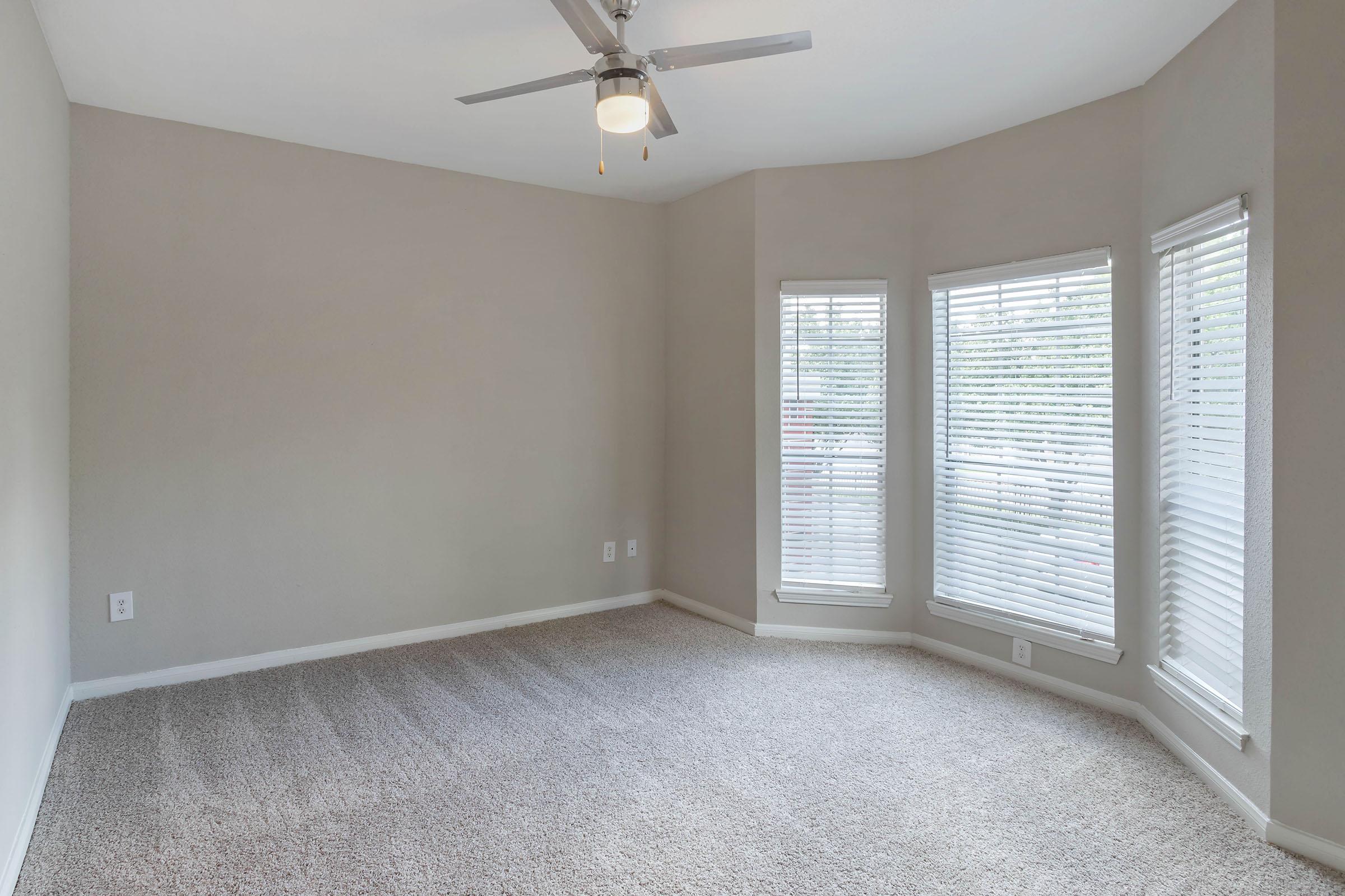 A spacious room with light gray walls and plush carpet. It features a ceiling fan and two large windows with white blinds, allowing natural light to fill the space. The room has a cozy, inviting atmosphere, ideal for various interior design options.