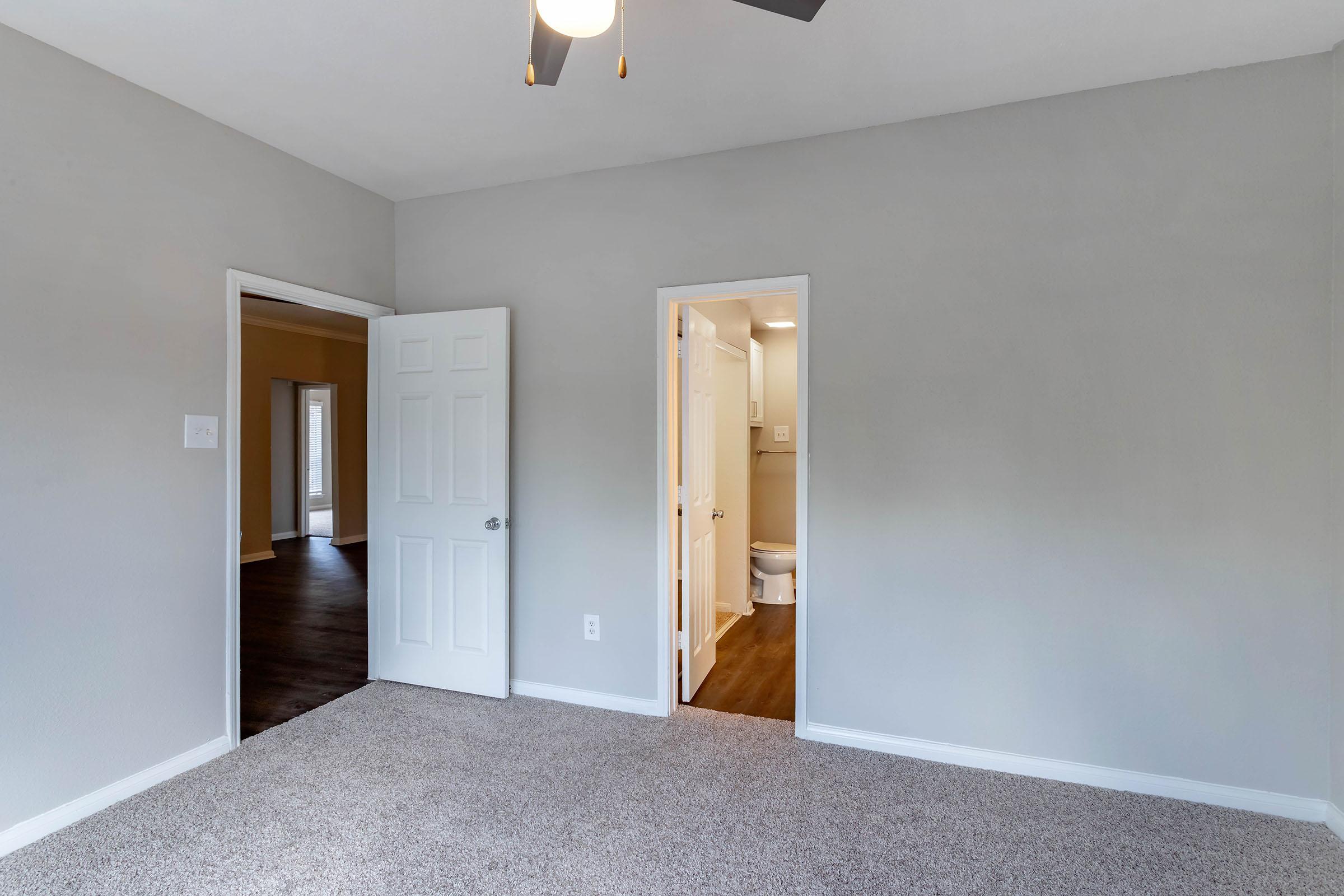 Interior view of a room featuring light gray walls and carpeted flooring. A ceiling fan is installed. A white door opens to an adjoining bathroom, while another door leads to a different area. Natural light enters from a window, enhancing the space's brightness and openness.