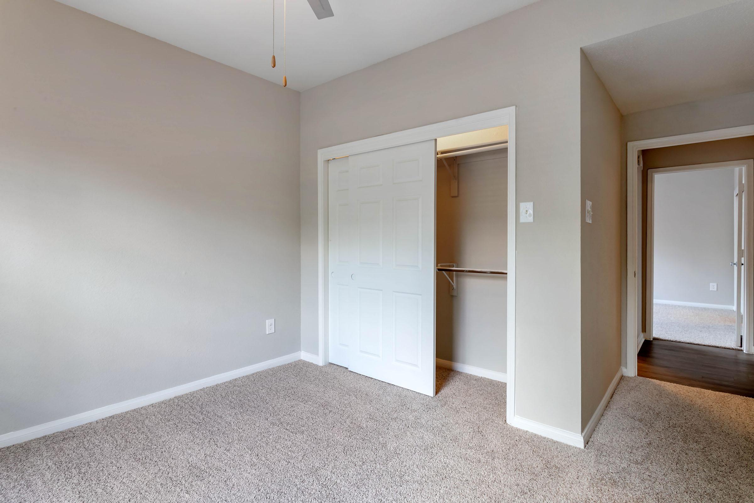 A bright, empty room featuring light gray walls and beige carpet. On the left, there is a closed white double door leading to a closet, and a ceiling fan above. A doorway on the right leads to another room, creating a sense of openness and potential for various uses.