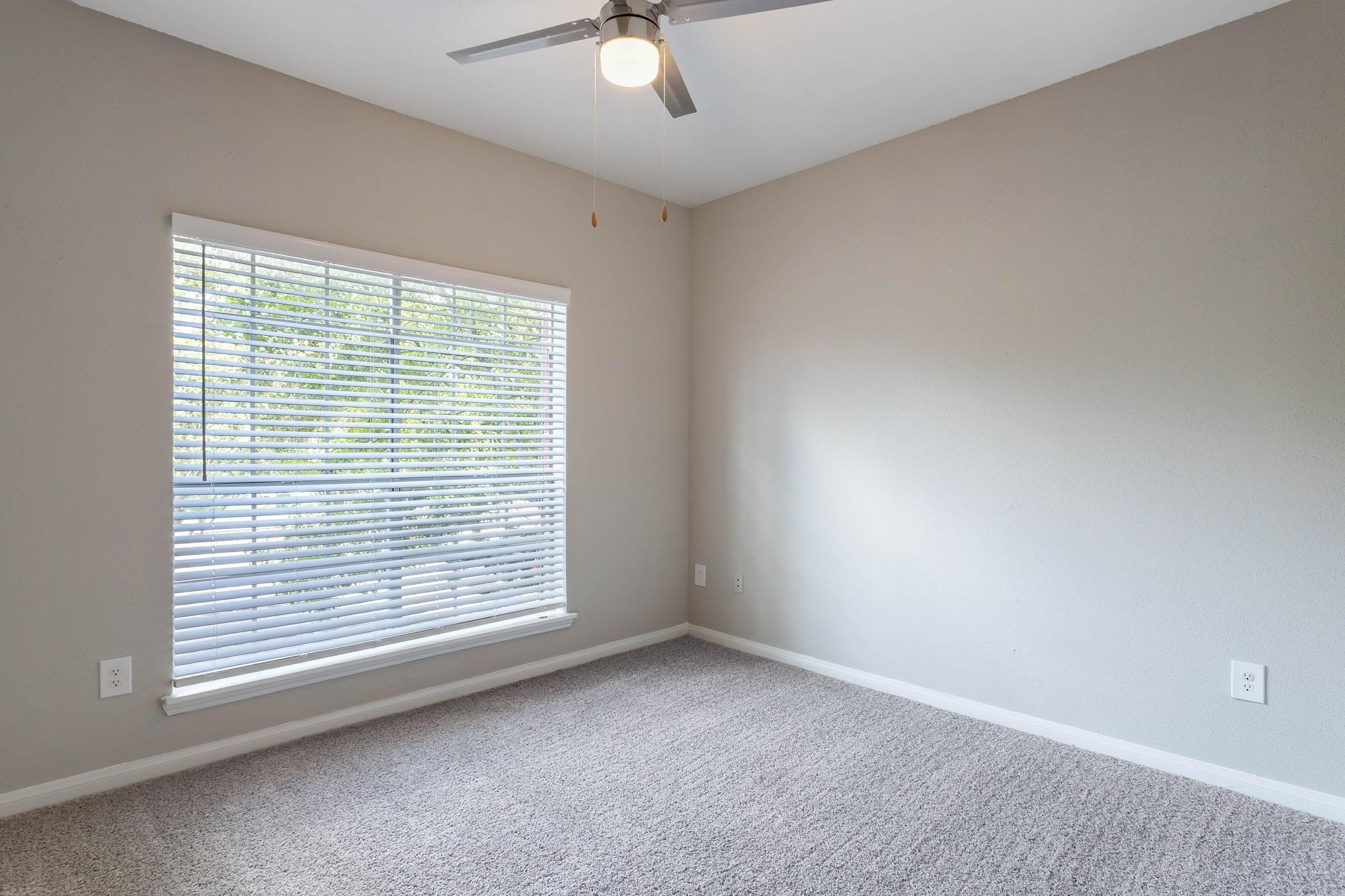 Empty room with light gray walls and carpeted flooring. A white window with horizontal blinds lets in natural light. A ceiling fan is mounted on the ceiling, adding a modern touch. The space is uncluttered, creating a clean and minimalistic atmosphere. No furniture is present in the room.