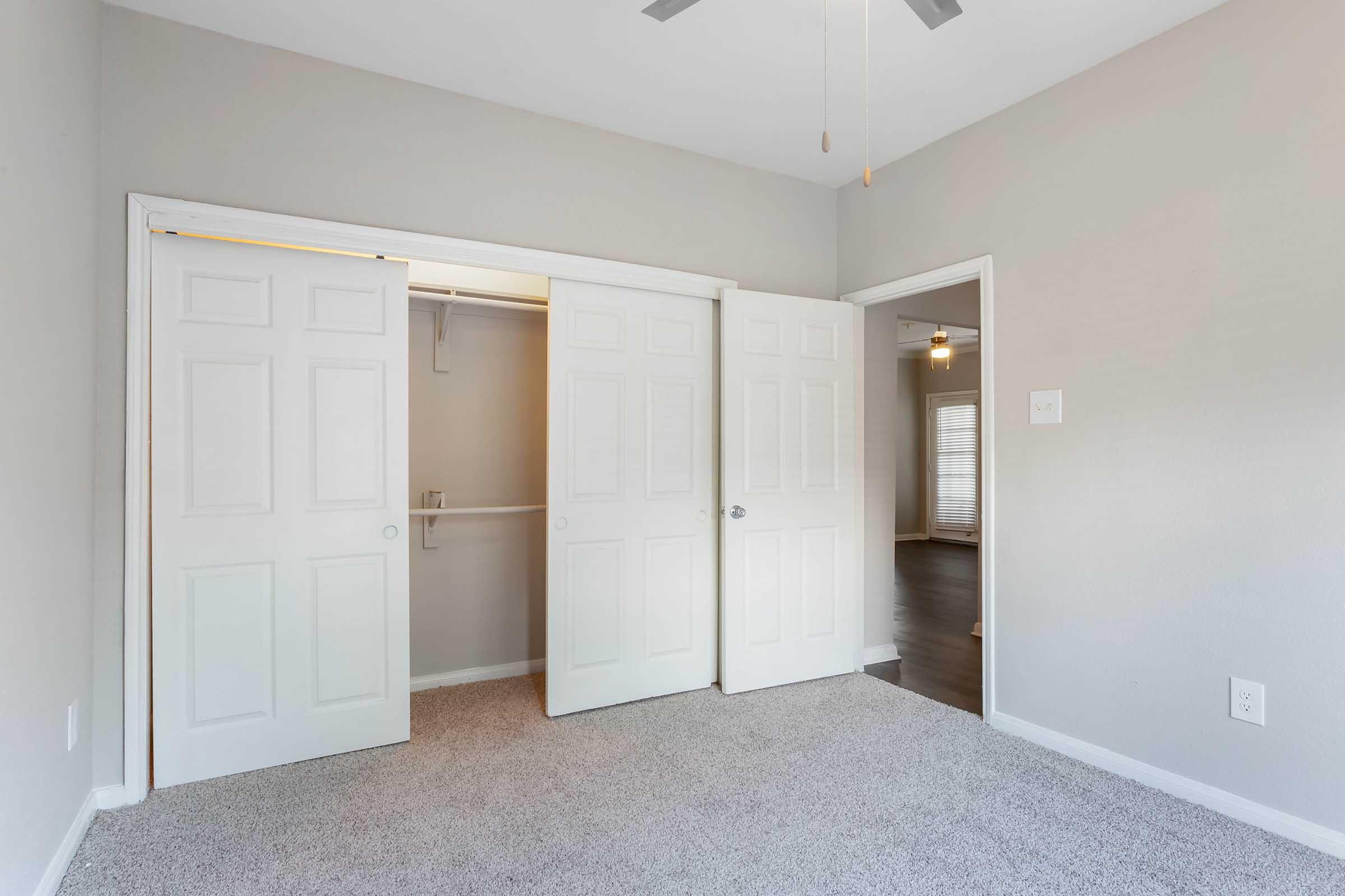 Empty bedroom with light-colored walls and carpeted floor. Two sets of white double doors open into a closet. A ceiling fan is visible above, and a doorway leads to another room in the background. Soft natural light streams in through a nearby window.