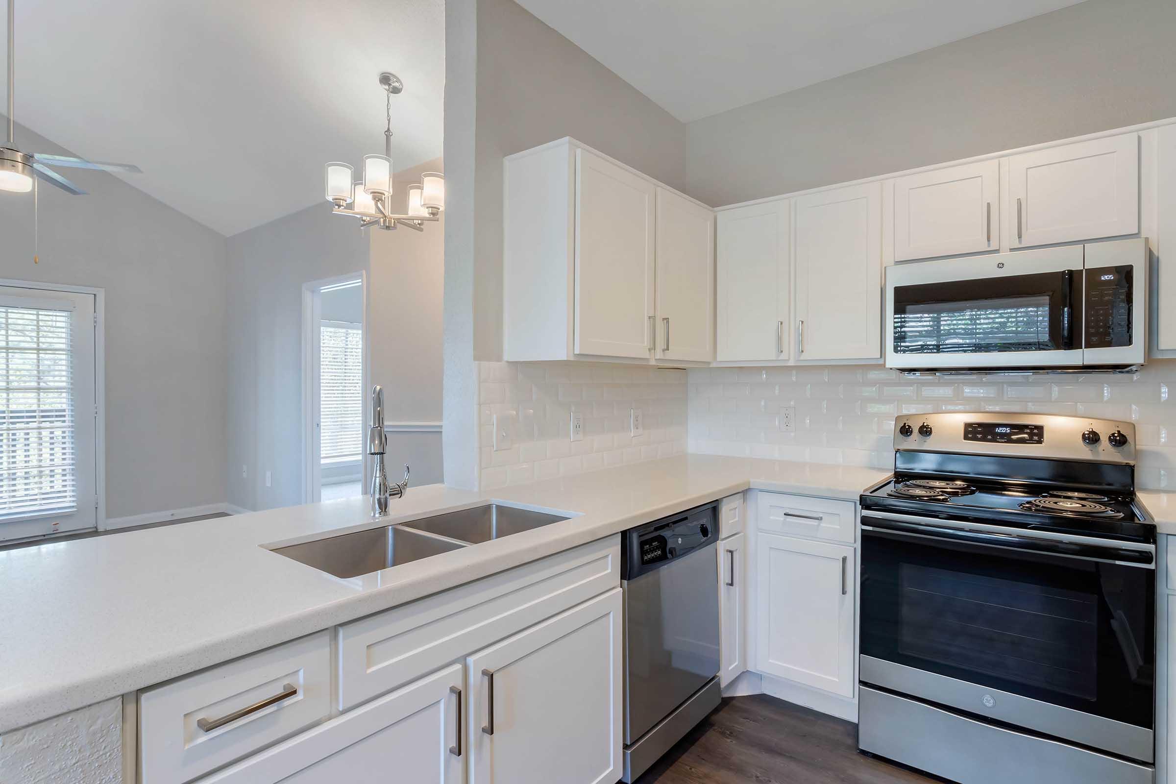 Modern kitchen featuring white cabinetry, stainless steel appliances including a microwave and stove, a dishwasher, and a double sink. The space has a light-colored countertop and walls, with large windows allowing natural light. A ceiling fan and stylish light fixture enhance the contemporary design.