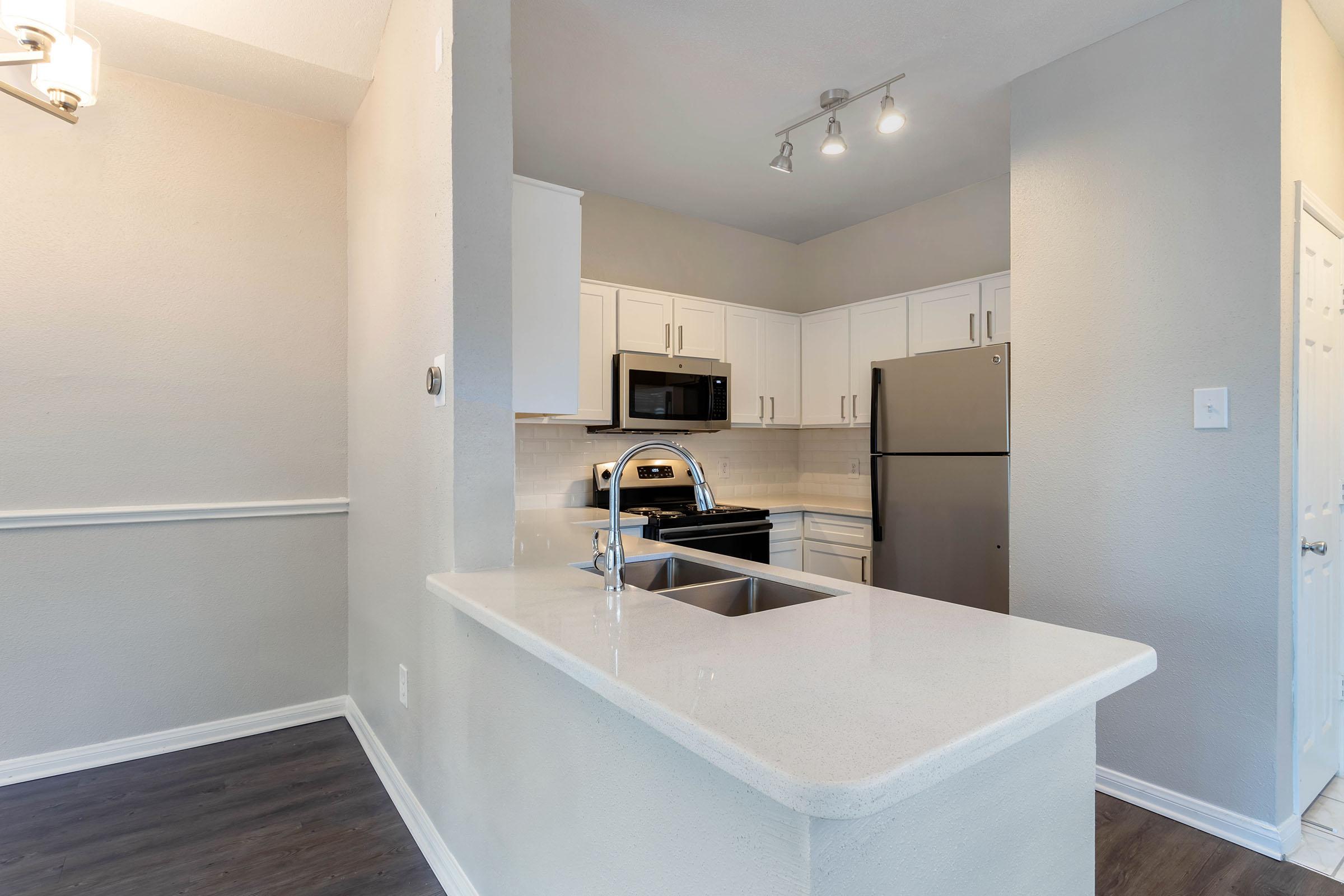 Modern kitchen interior featuring white cabinetry, stainless steel appliances, and a granite countertop with a double sink. The space includes overhead lighting, a built-in microwave, and a refrigerator, with a light-colored backsplash and dark laminate flooring. Overall, the design is bright and inviting.
